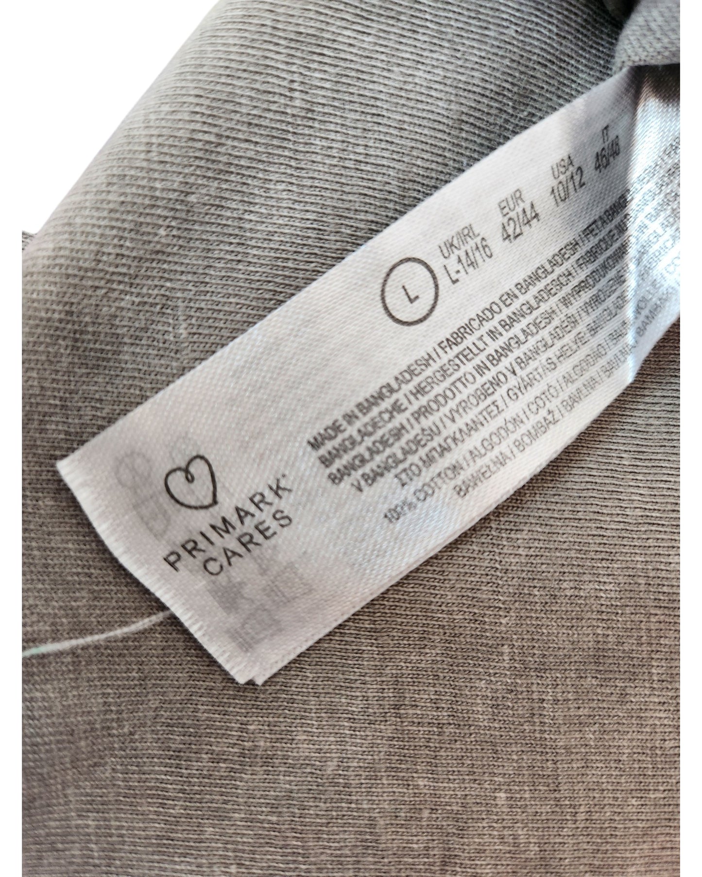 A close-up of a Primark label on a grey Upcycled Breastfeeding & Pumping Nirvana Band t-shirt (L), displaying “PRIMARK CARES” with heart logo, eco-friendly message, and size info: UK/IRL 14/16, EUR 42/44, USA 10/12.
