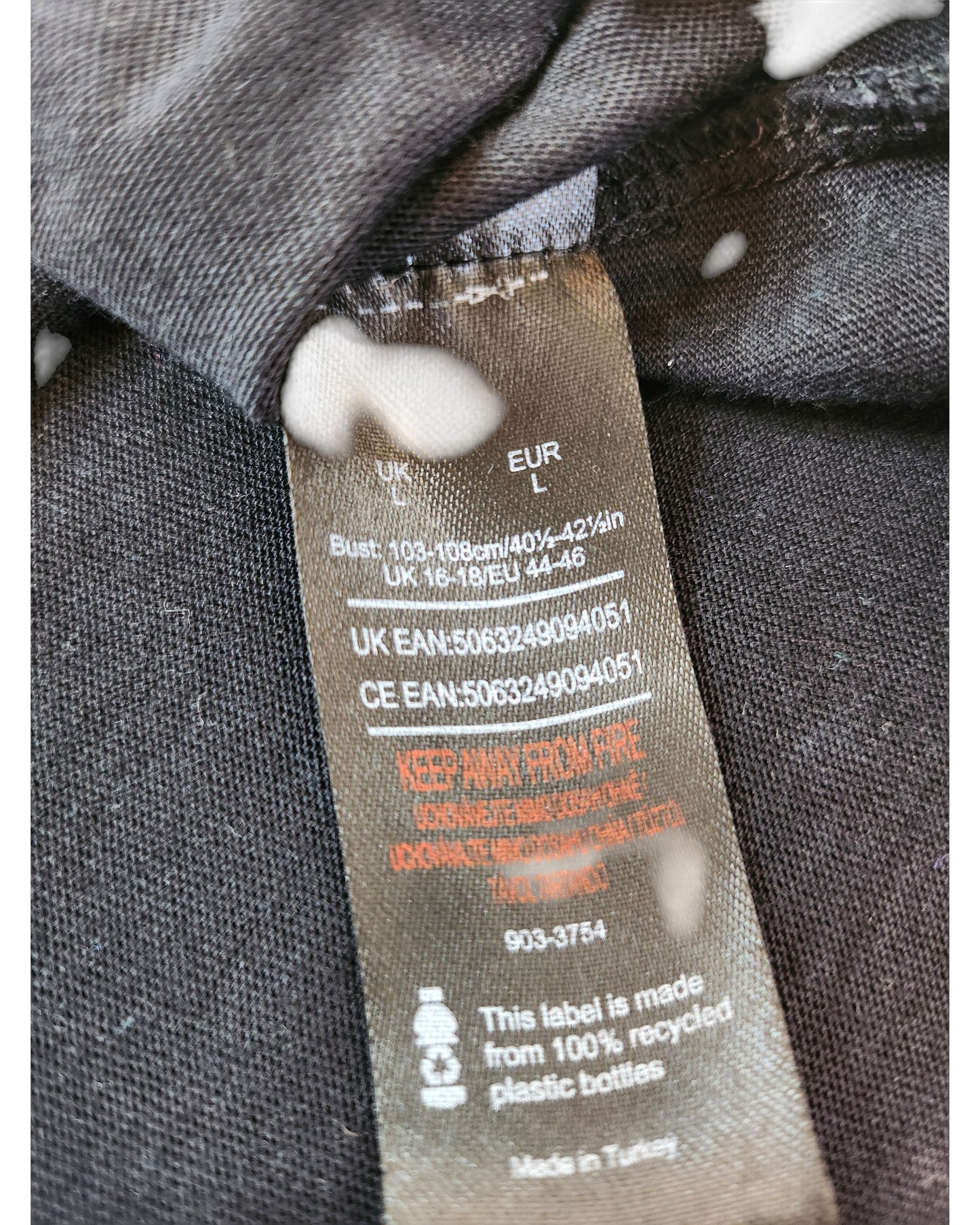 A close-up of a clothing tag on a black Def Leppard Upcycled Breastfeeding & Pumping Band t-shirt (L) with white spots. The partially folded tag lists sizing, care info, and notes it’s made from 100% recycled plastic bottles.