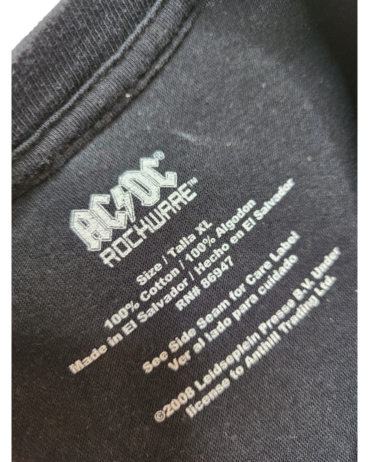 Close-up of an AC/DC Upcycled Breastfeeding & Pumping Back in Black Band t-shirt tag, size XL, 100% cotton, made in El Salvador—ideal for fans seeking unique, comfy nursing apparel with brand and copyright info.