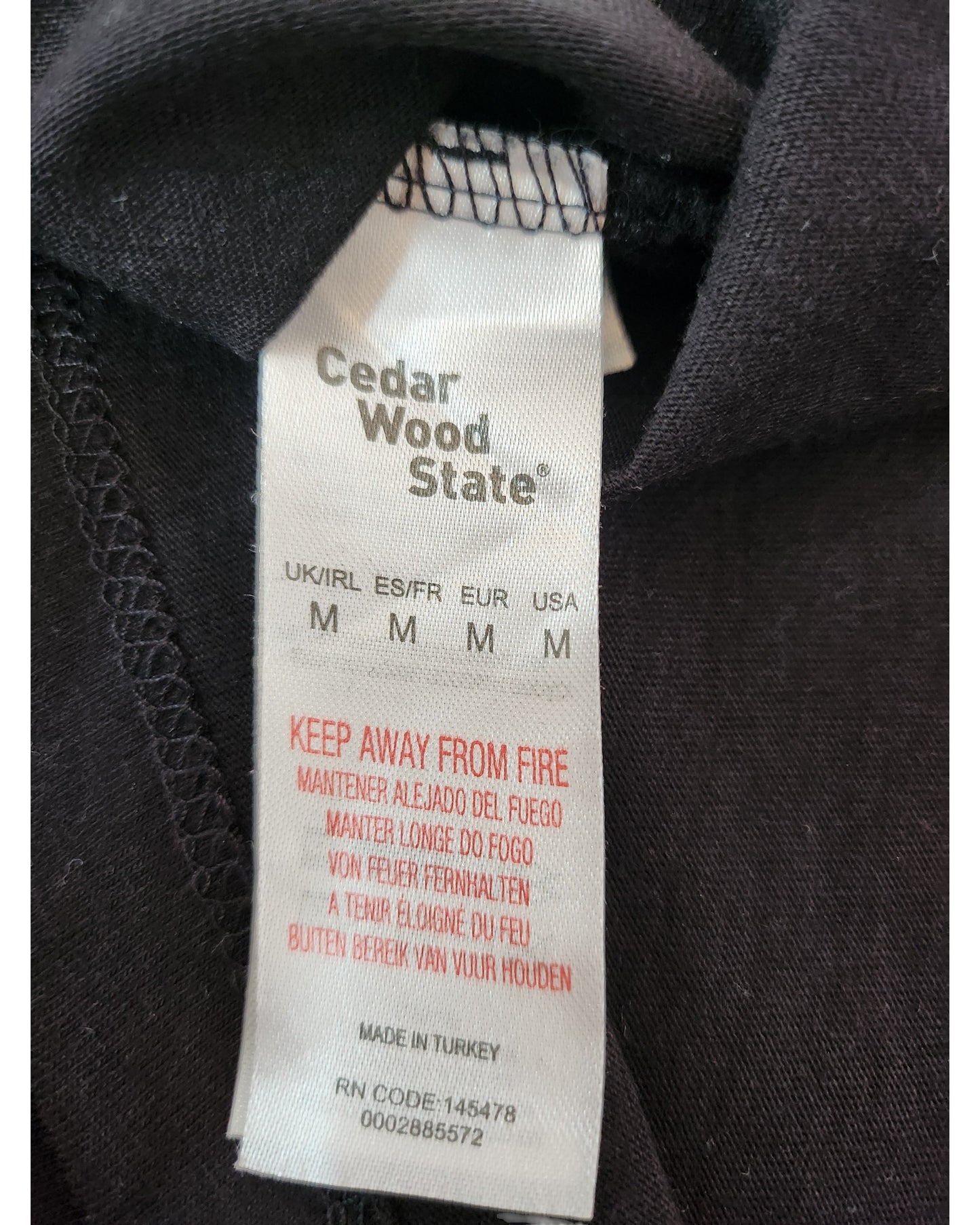 A close-up of a Cedar Wood State label on an M-size Upcycled Breastfeeding & Pumping Space Invaders t-shirt, featuring a multilingual Keep Away from Fire warning and noting upcycled origin. Made in Turkey.