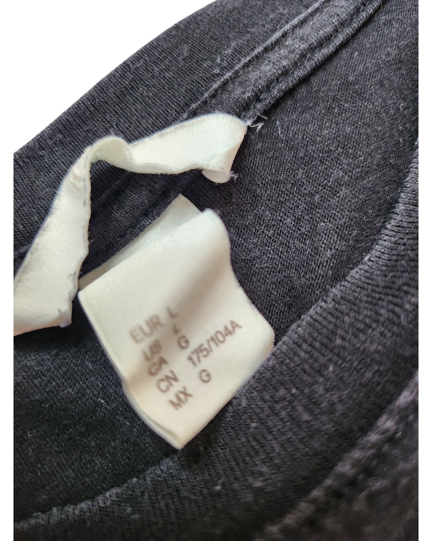 Close-up of a white H&M clothing tag on an upcycled black floral embroidered breastfeeding & pumping t-shirt (L), showing size info for EUR, USA, GB, CN, MX as G/large. Tag is partially folded—a perfect find for preloved fashion lovers.