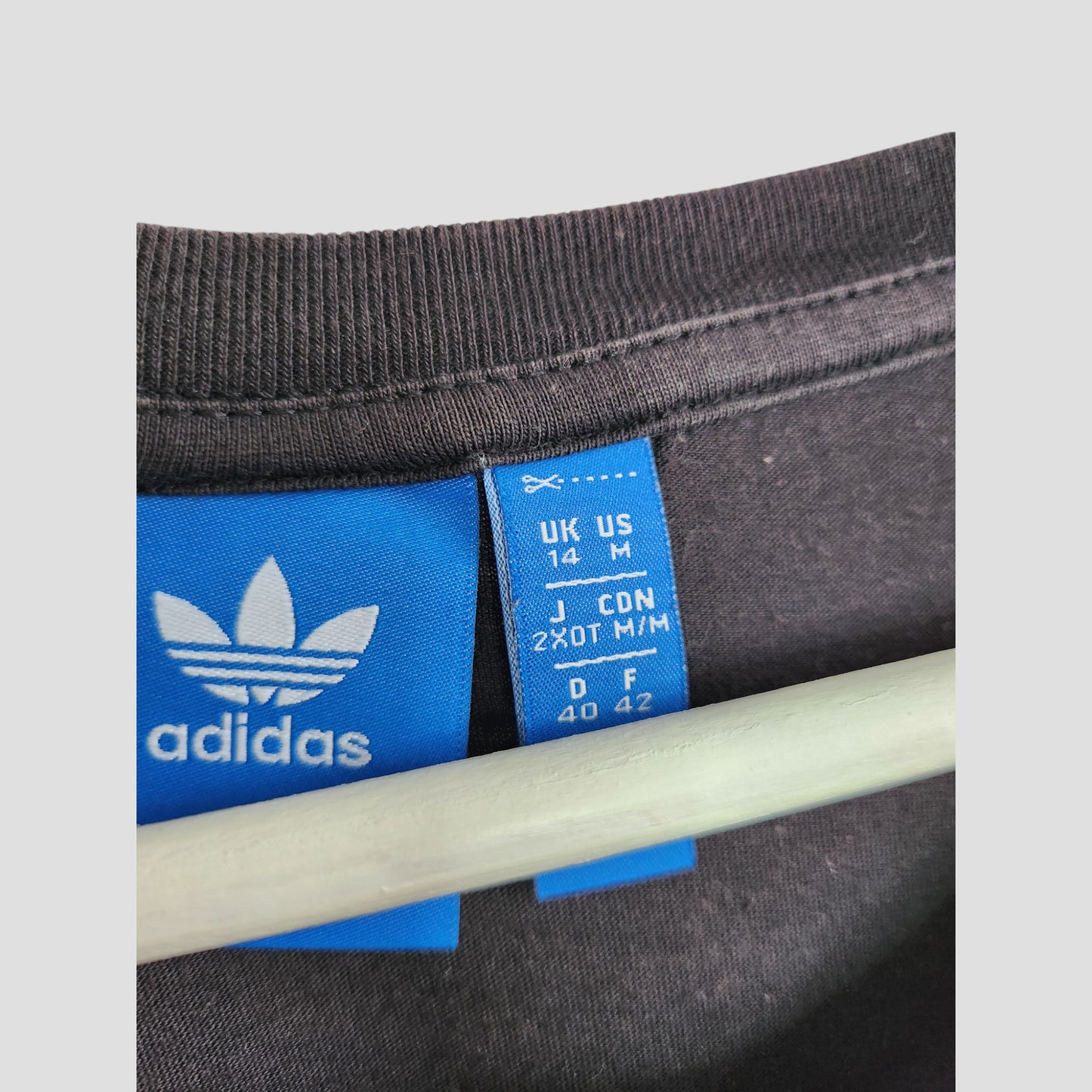 Close-up of a preloved Adidas tag with sizes UK14/US M/D40/F42, attached to an upcycled black/white breastfeeding & pumping t-shirt (UK14) by Adidas, partially covered by a hanger.