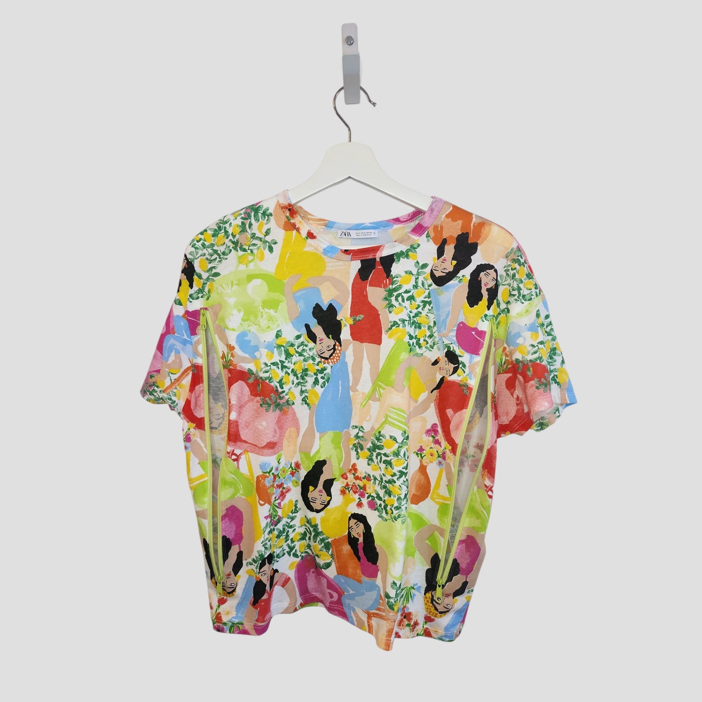 The ZARA Upcycled Breastfeeding & Pumping Girls Printed t-shirt (S, 100254) features a vibrant abstract pattern with women in dresses and florals. Eco-friendly, its shown on a white hanger against a plain light background.