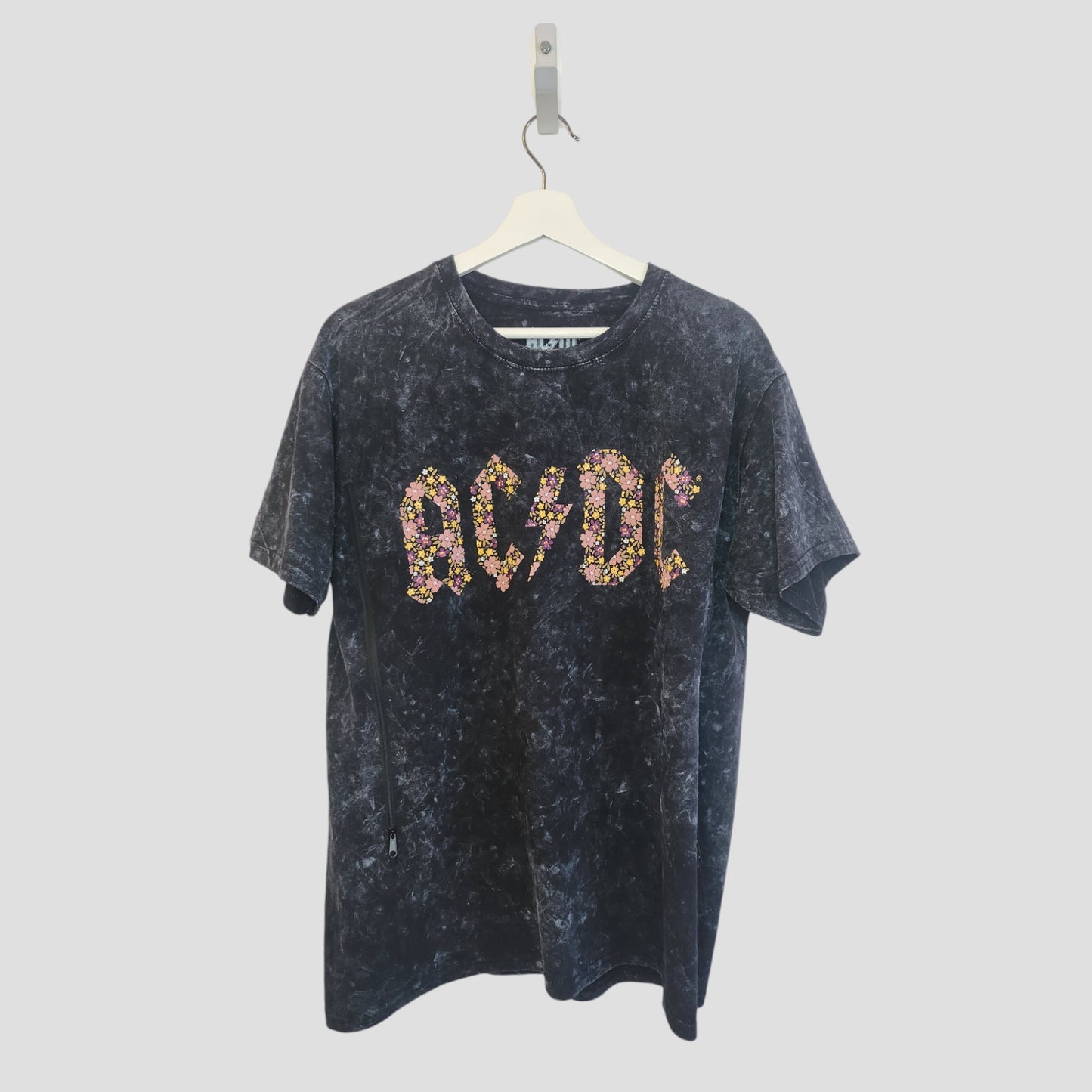 An upcycled black floral AC/DC band t-shirt (L) for breastfeeding and pumping hangs on a white hanger against a plain background, featuring the AC/DC logo in a colorful floral pattern.