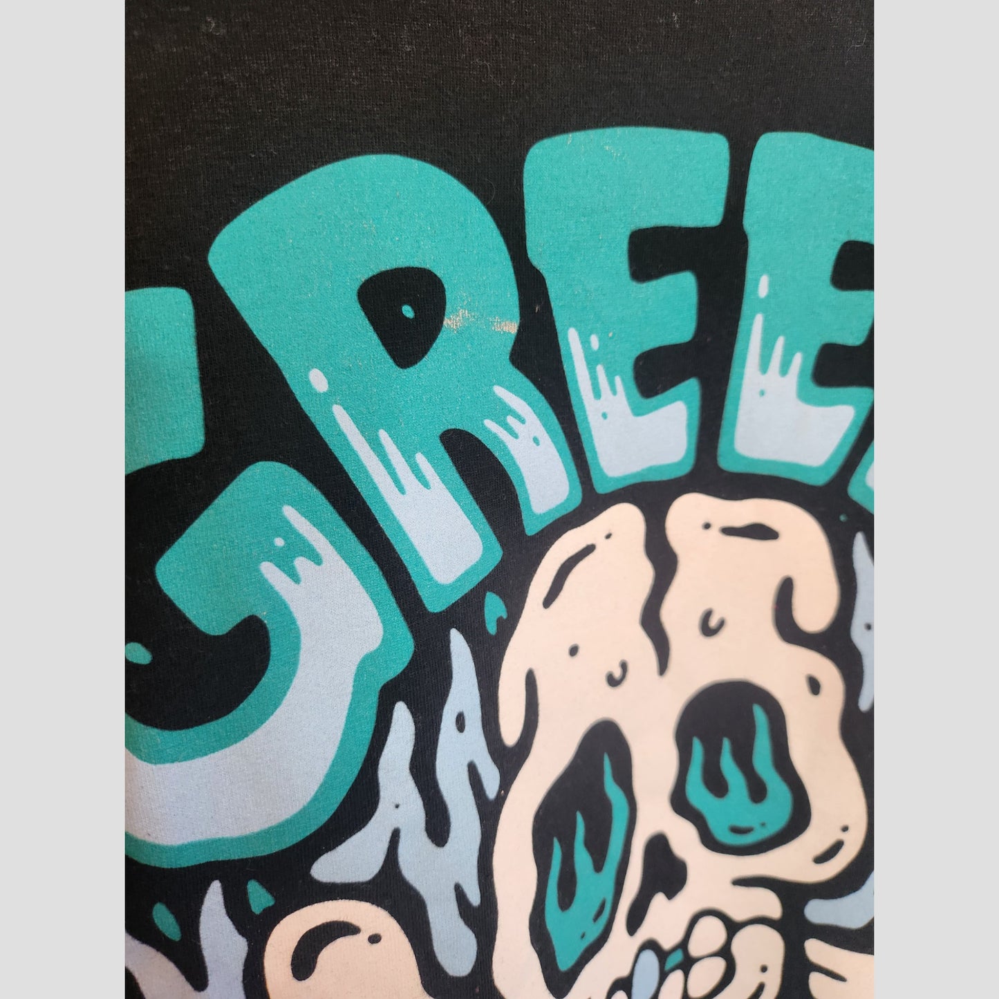 Partial view of bold turquoise GREE text above an illustrated teal and black skull on black, inspired by upcycled breastfeeding clothing. Product: Upcycled Breastfeeding & Pumping Black Skull Green Day Band t-shirt XL by Gildan.