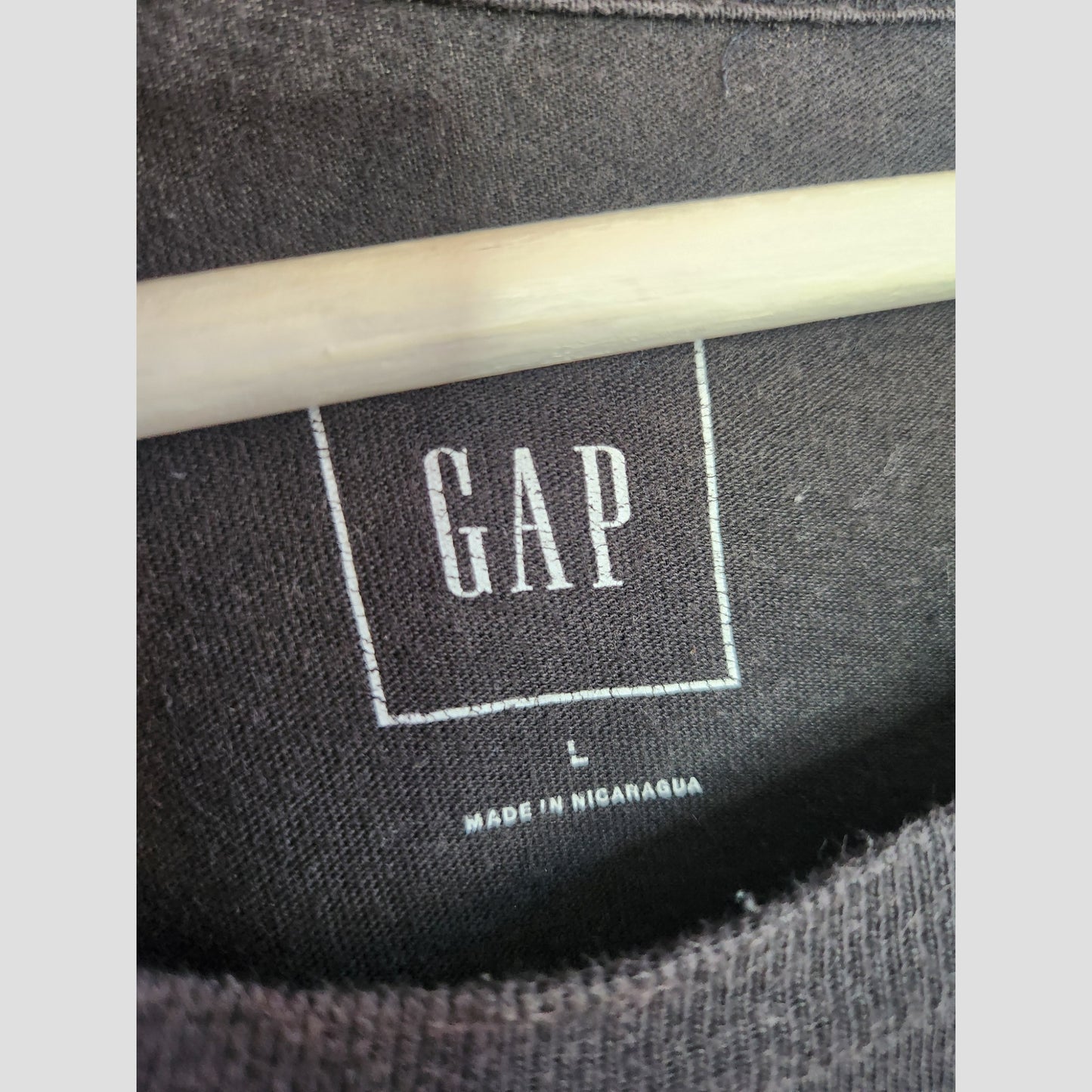 Close-up of a black GAP T-shirt label with GAP in white letters, size L, and MADE IN NICARAGUA printed below. Wooden hanger shown. Product: Upcycled Breastfeeding & Pumping Metallica GAP Band t-shirt L (100085) by GAP.