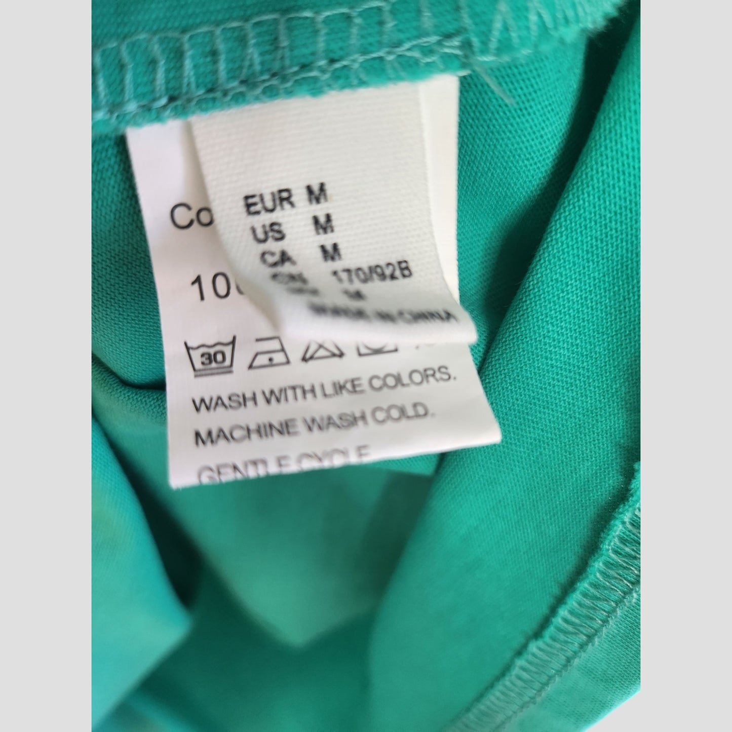 A close-up of the care label on our Upcycled Breastfeeding & Pumping Green White Fox t-shirt (M, 100266) displays size info (EUR M, US M, CA M) and advises cold gentle wash with like colors.