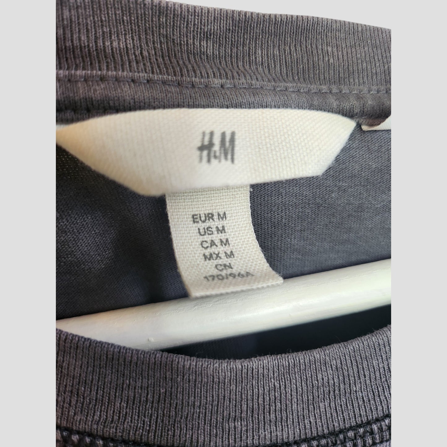 Close-up of an H&M tag on an Upcycled Breastfeeding & Pumping Grey Pink Floyd Rainbow Band t-shirt (M, 100256), shown on eco-friendly maternity wear hanging on a white hanger.