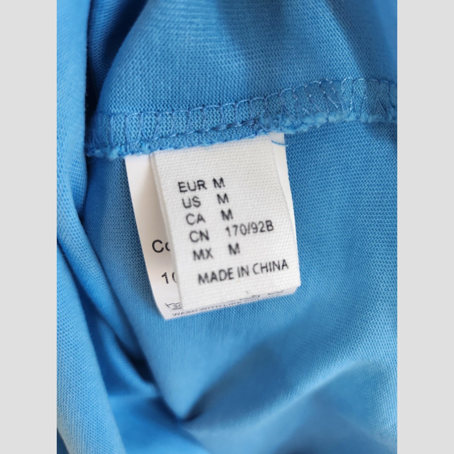 Close-up of a tag on the Upcycled Breastfeeding (Nursing) & Pumping Blue White Fox t-shirt M (100265), showing size Medium for various regions and Made in China—great for those seeking preloved nursing wear. Brand: Unknown.