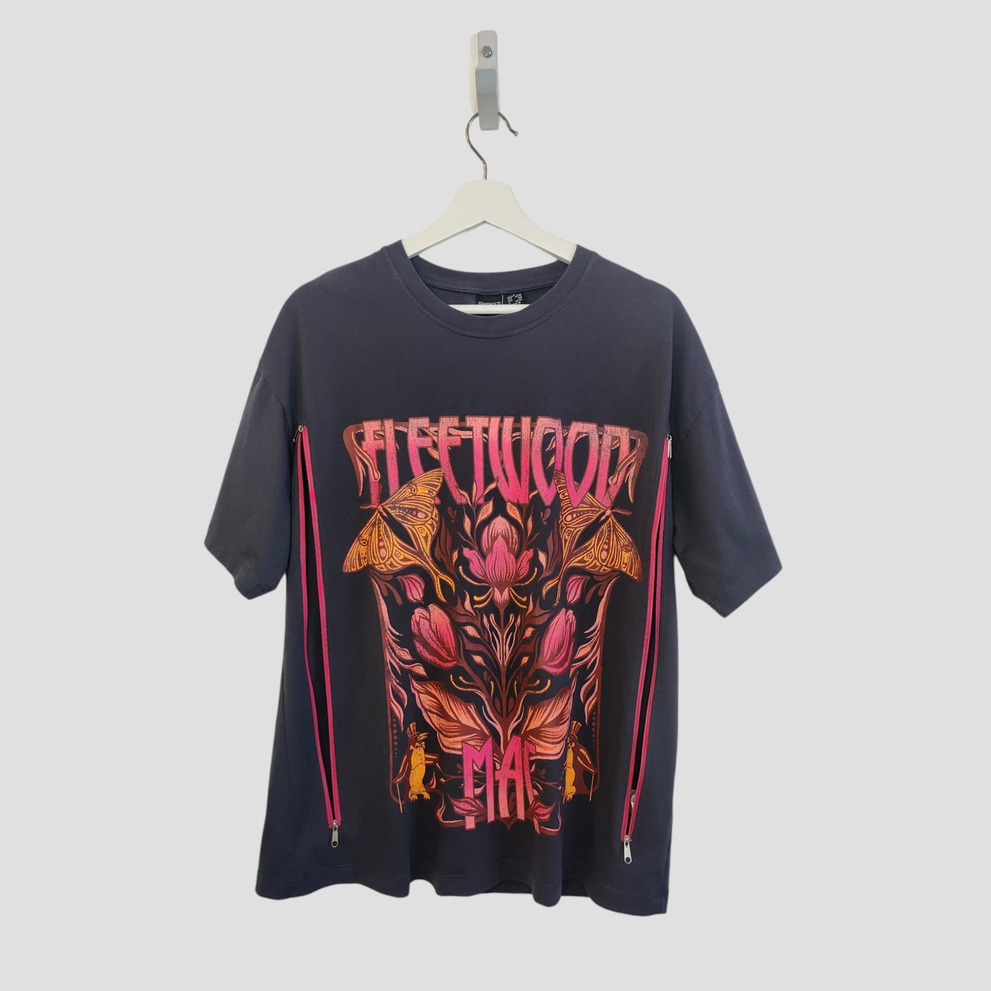 A grey floral Fleetwood Mac band t-shirt by Primark hangs on a white hanger. Upcycled for breastfeeding and pumping, it features bold pink/orange text, vibrant floral/moth graphics, and pink side zippers. Size S (UK10/12).