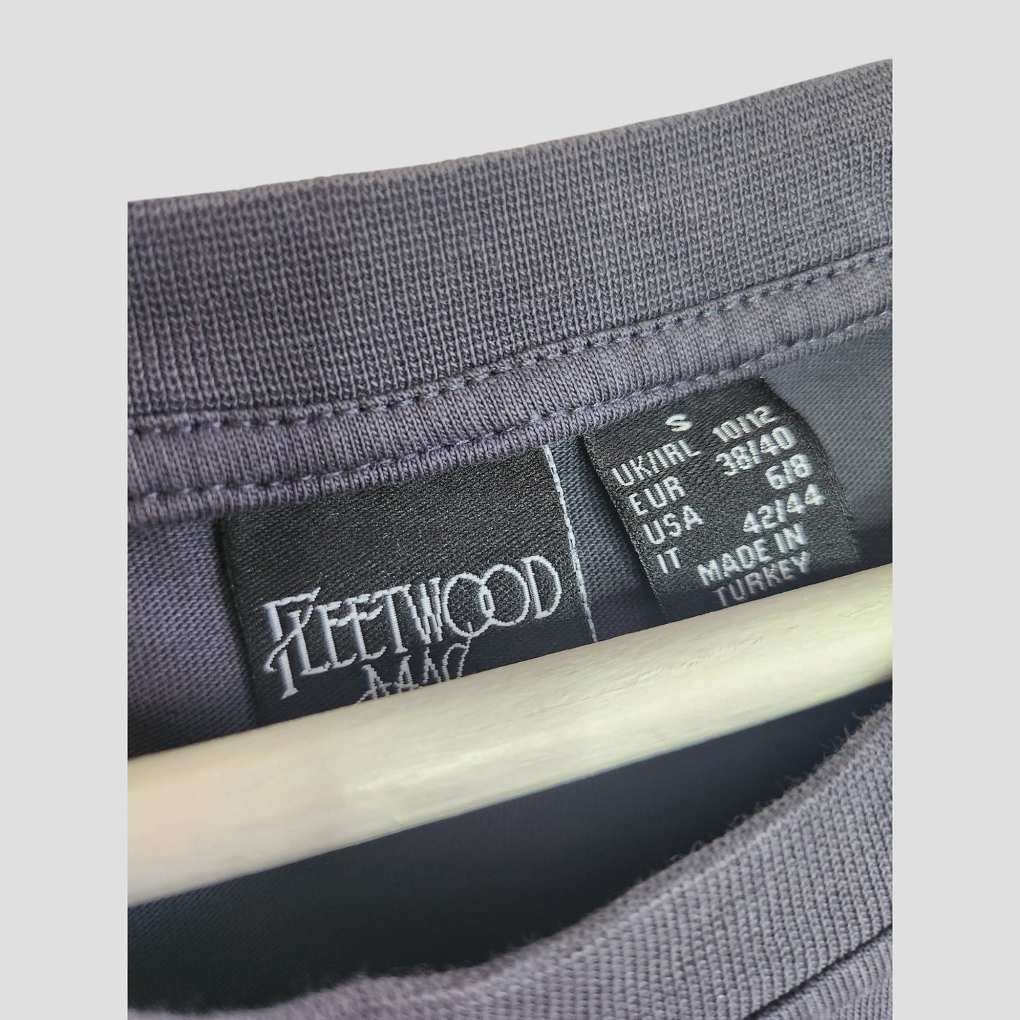 Close-up of a black Primark shirt collar with two tags—one featuring Fleetwood Mac, the other with sizing and Made in Turkey details. The hanger shows this Upcycled Breastfeeding & Pumping Grey Floral Fleetwood Mac Band tee S (UK10/12).