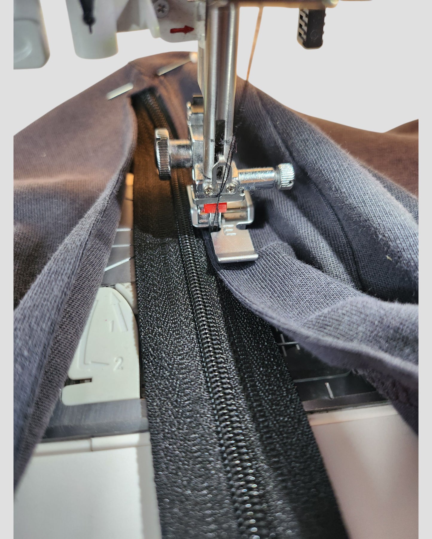 A close-up of The Original Sew-On Zip Kit for Breastfeeding & Pumping Access by touched out mama shows a sewing machine stitching a black zipper between two fabric pieces—ideal for DIY nursing projects. Needle and presser foot are in focus.