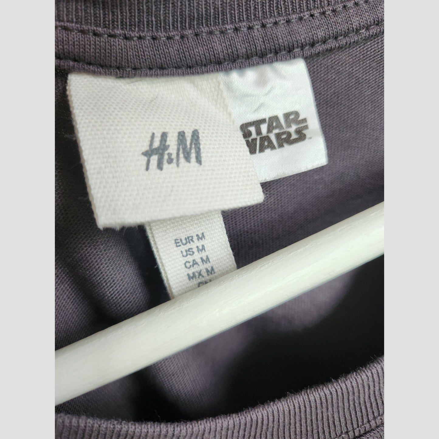 Close-up of H&M Upcycled Breastfeeding & Pumping Grey Star Wars t-shirt (M) tags, highlighting medium size for Europe, US, Canada, and Mexico. Eco-friendly item on a white hanger.