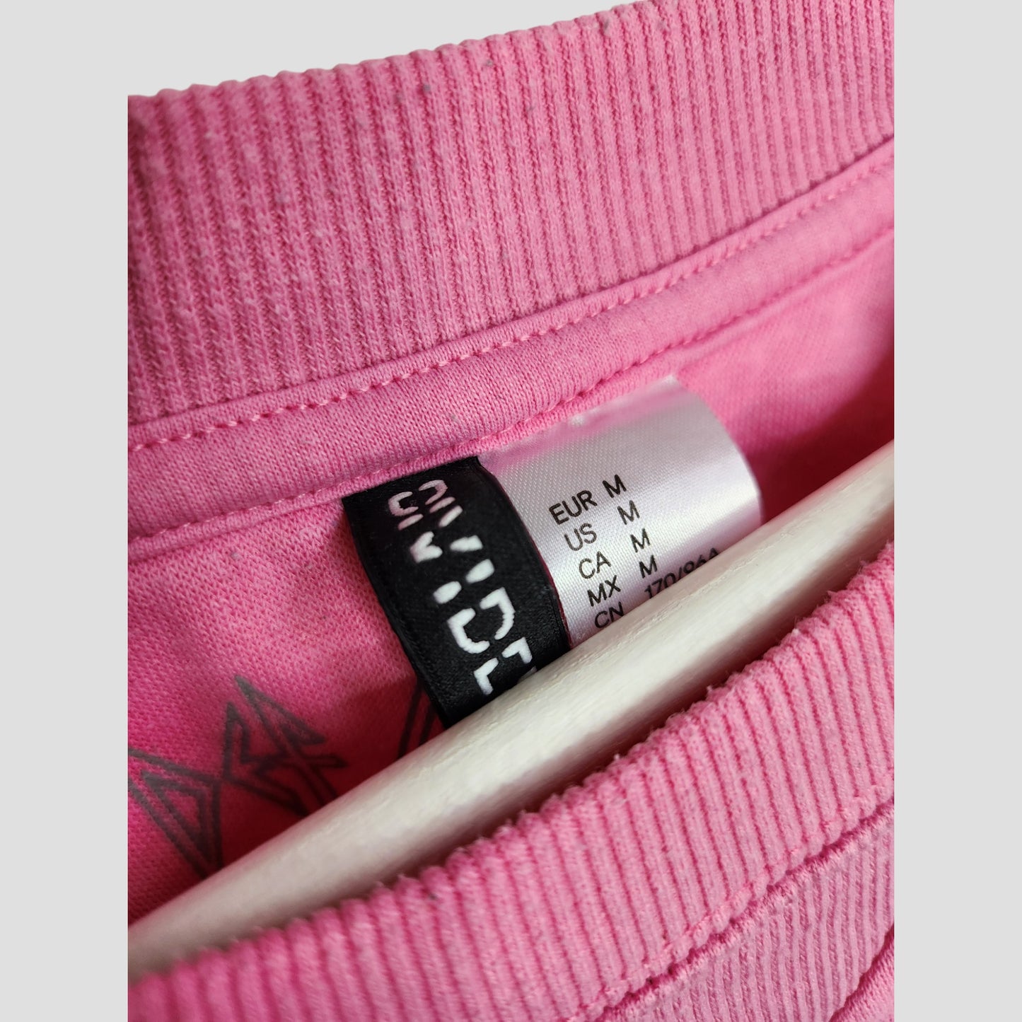 Close-up of a pink H&M Upcycled Breastfeeding & Pumping Def Leppard Band Sweatshirt (M, 100131) collar with regional size tag and brand label, emphasizing its eco-friendly, pre-loved origins. A hanger is visible beneath.