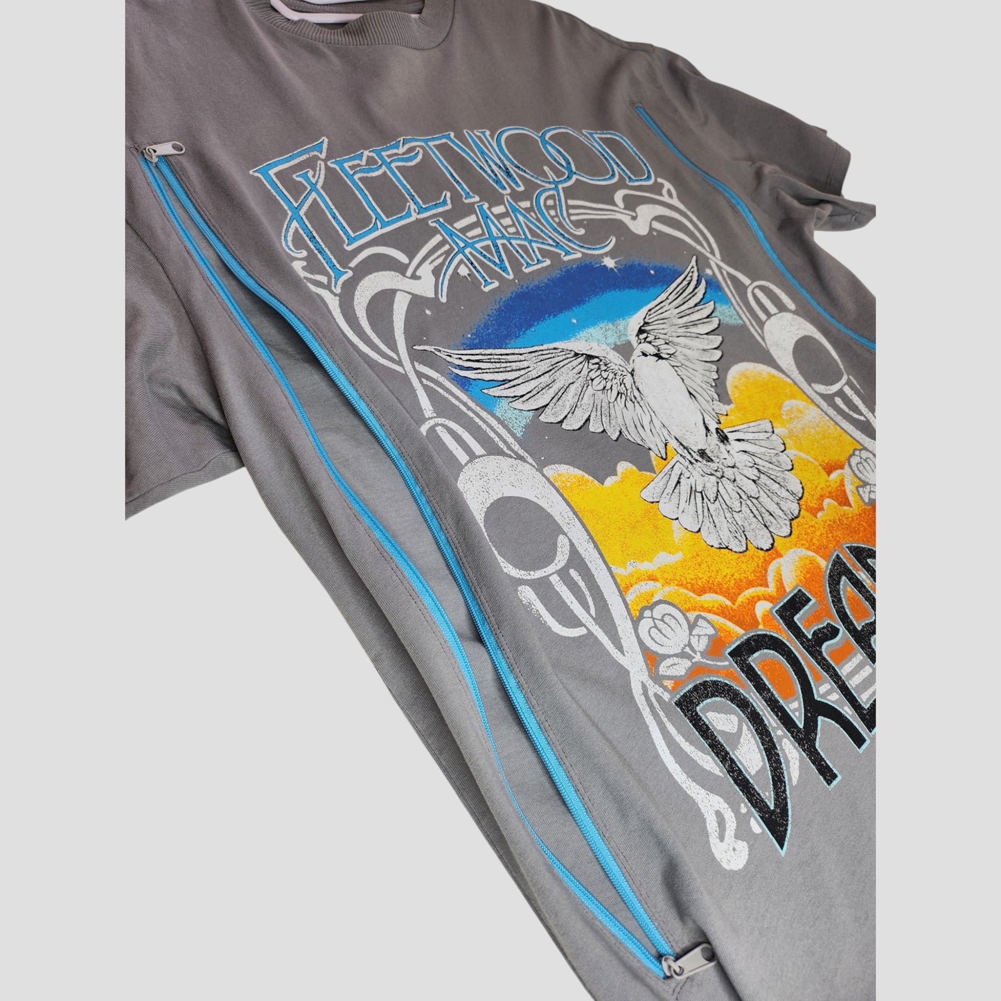 The H&M Upcycled Fleetwood Mac Dreams Band t-shirt (S) is transformed for nursing & pumping, featuring a white dove, blue sky, orange clouds, “DREAMS” text, and vertical side zippers—perfect eco-friendly maternity wear.