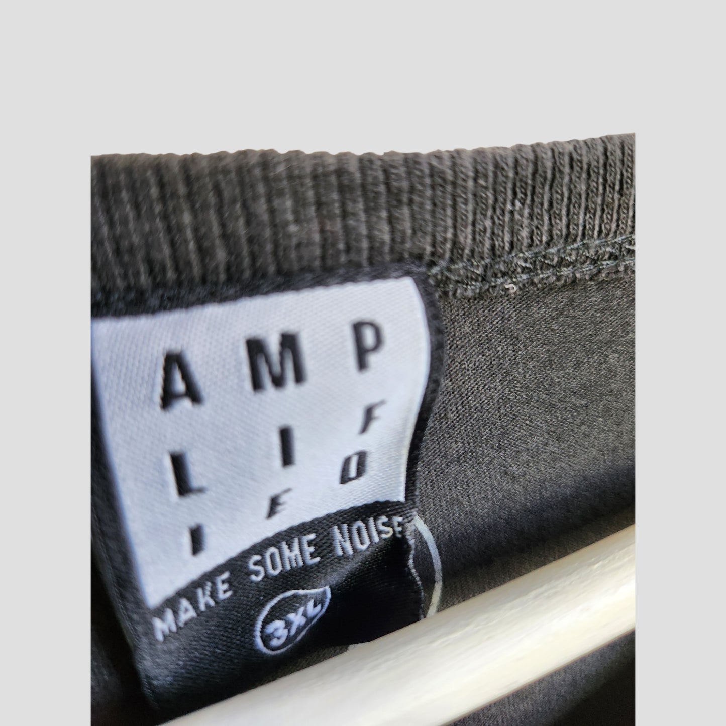 Close-up of a black clothing tag inside an upcycled Amplified breastfeeding & pumping grey Foo Fighters band t-shirt (3XL), showing "AMPLIFE, MAKE SOME NOISE," and the size in white on black.
