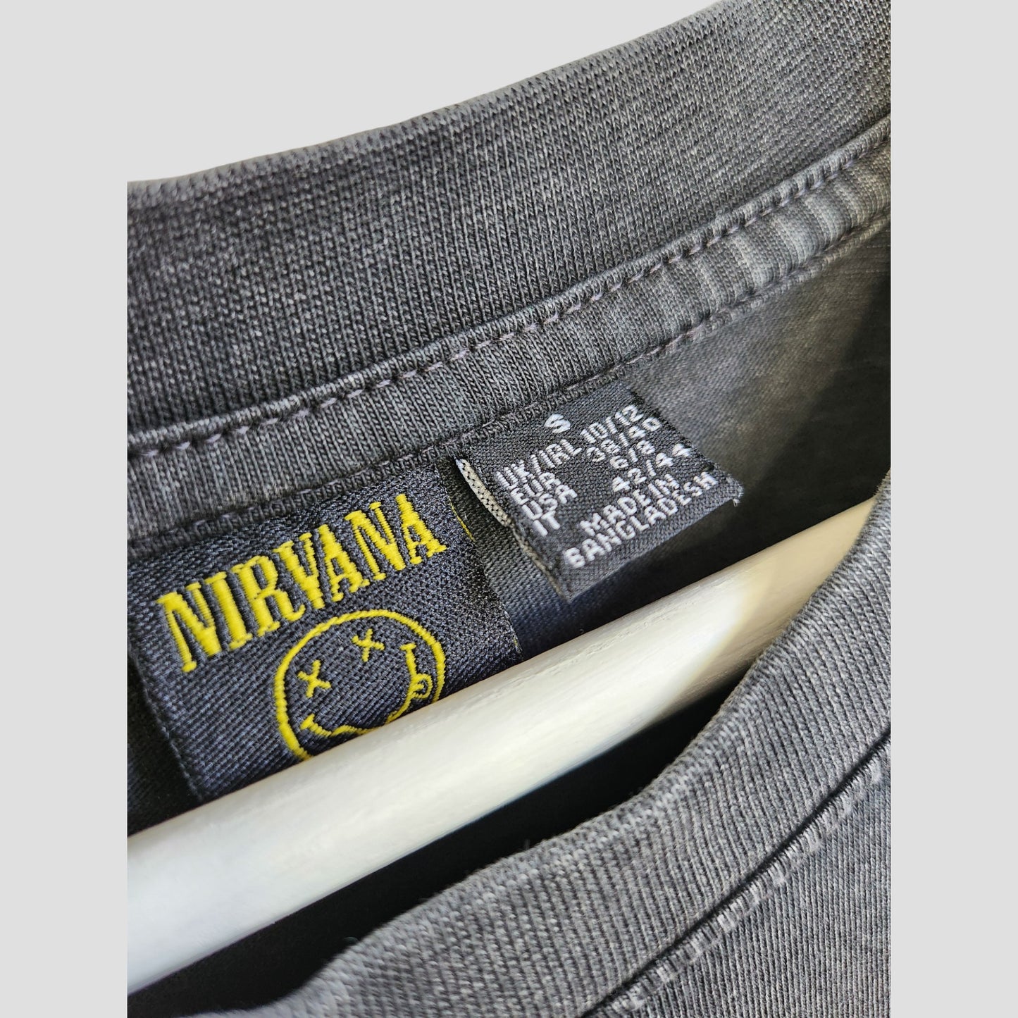Close-up of a dark grey Primark Nirvana band T-shirt collar with floral design, yellow smiley label, and black care tag—an upcycled, eco-friendly oversized breastfeeding/pumping top in size S (UK10/12).