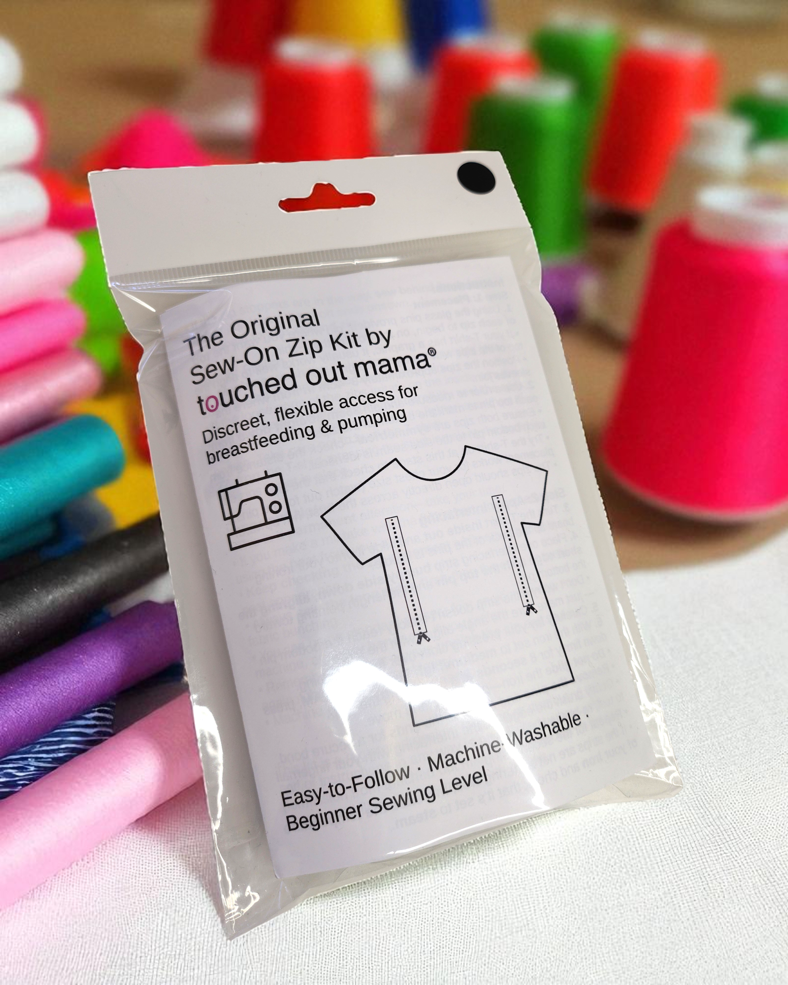 A packet of The Original Sew-On Zip Kit by touched out mama sits on a table with colorful thread. This kit helps upcycle t-shirts with zippers and instructions for breastfeeding access, supporting reduced textile waste.