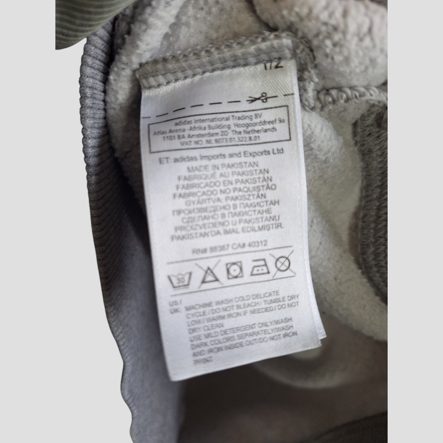 A close-up of the care label on a grey/white upcycled Adidas breastfeeding & pumping sweatshirt (L, UK14), showing washing instructions, materials, manufacturing details in several languages, and indicating it was made in Pakistan.