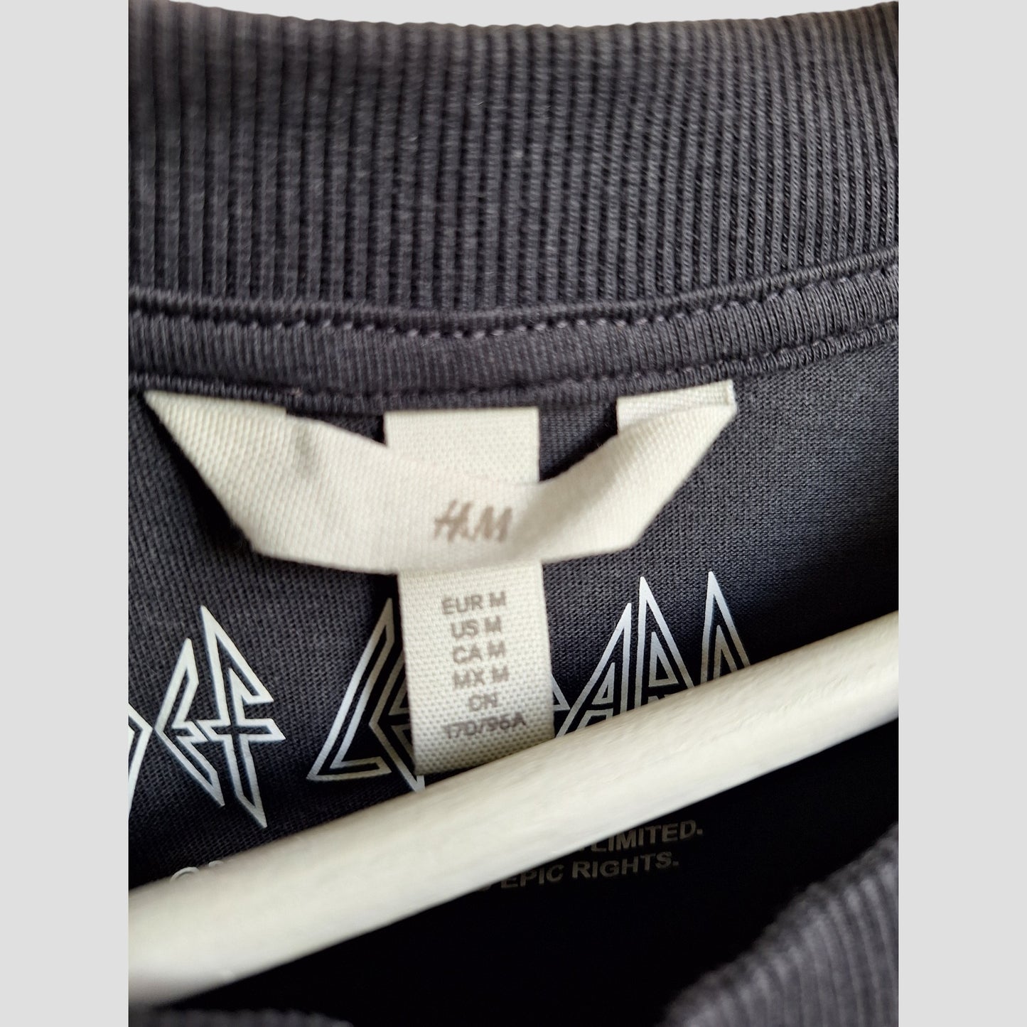 Close-up of an H&M tag sewn into the Upcycled Breastfeeding & Pumping Grey Def Leppard Band t-shirt M (100370), showing size markers (EUR/US/CA/MX M, CN 170/96A) with the shirt’s graphic partly visible.