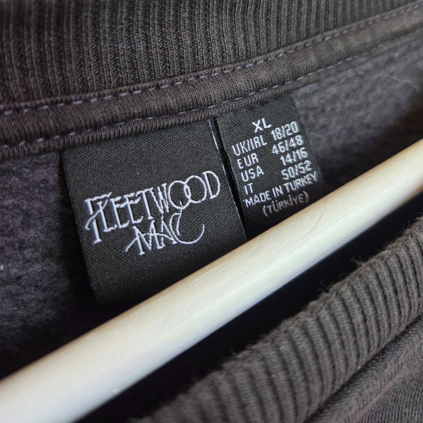Close-up of a black tag on a dark gray Primark Upcycled Breastfeeding & Pumping Floral Fleetwood Mac Band Sweatshirt XL (UK18/20), showing size info for UK, EU, USA. Eco-friendly item made in Turkey, hanging on a white hanger.