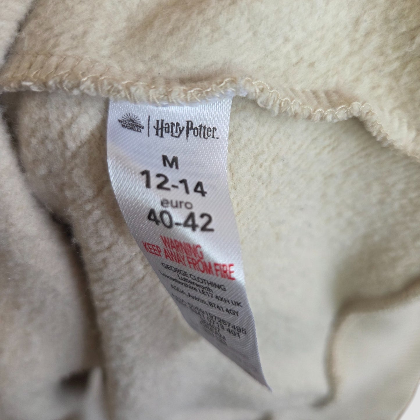 A close-up of a clothing tag on beige fabric shows the George Harry Potter logo, size M (UK12/14), and a red fire warning—ideal for fans seeking eco-friendly, upcycled breastfeeding or pumping sweatshirts.