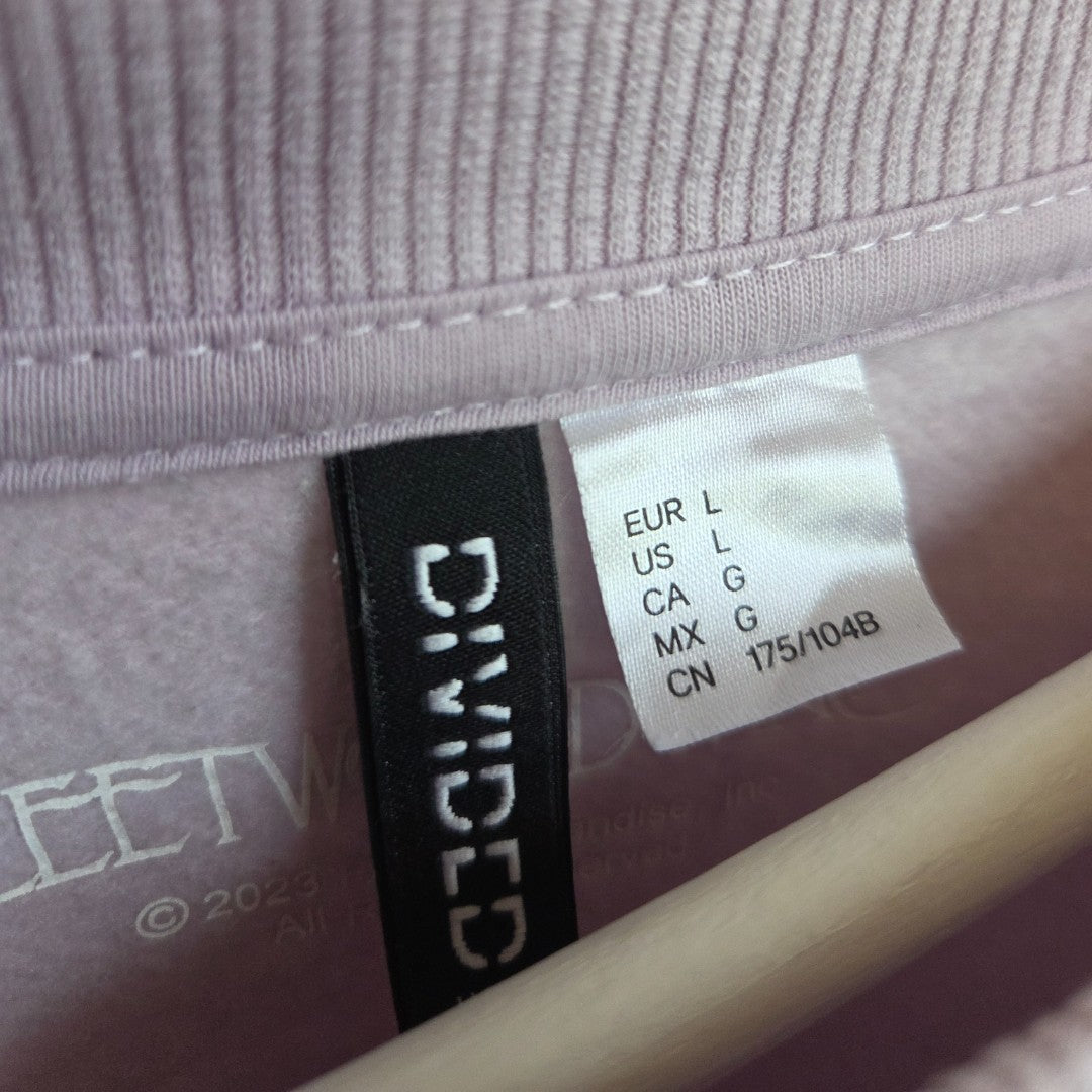 A close-up of clothing tags inside a lilac Upcycled Breastfeeding & Pumping Fleetwood Mac Band Sweatshirt (size L), showing regional size info and the H&M brand on eco-friendly, preloved apparel.