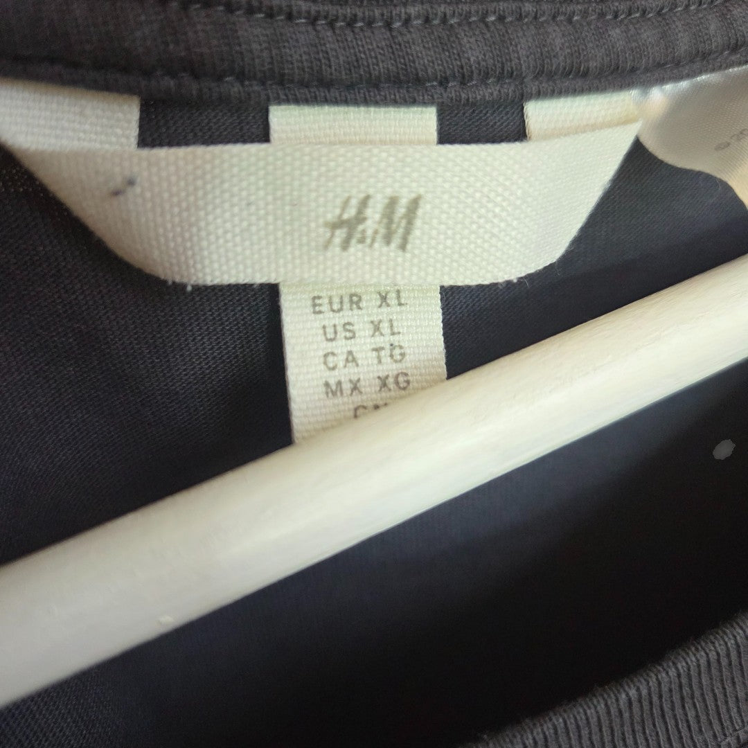 A close-up of an H&M tag on the Upcycled Breastfeeding & Pumping Grey White Nirvana Angel Band t-shirt XL (100460) shows size info: EUR XL, US XL, CA TG, MX XG. The dark garment hangs on a white hanger.