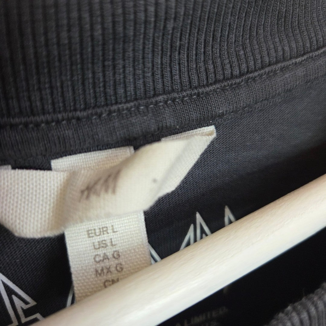 A close-up of an upcycled grey Def Leppard breastfeeding and pumping t-shirt by H&M, size L (100384), with a visible white H&M tag displaying size details for multiple regions, hanging on a light wood hanger.