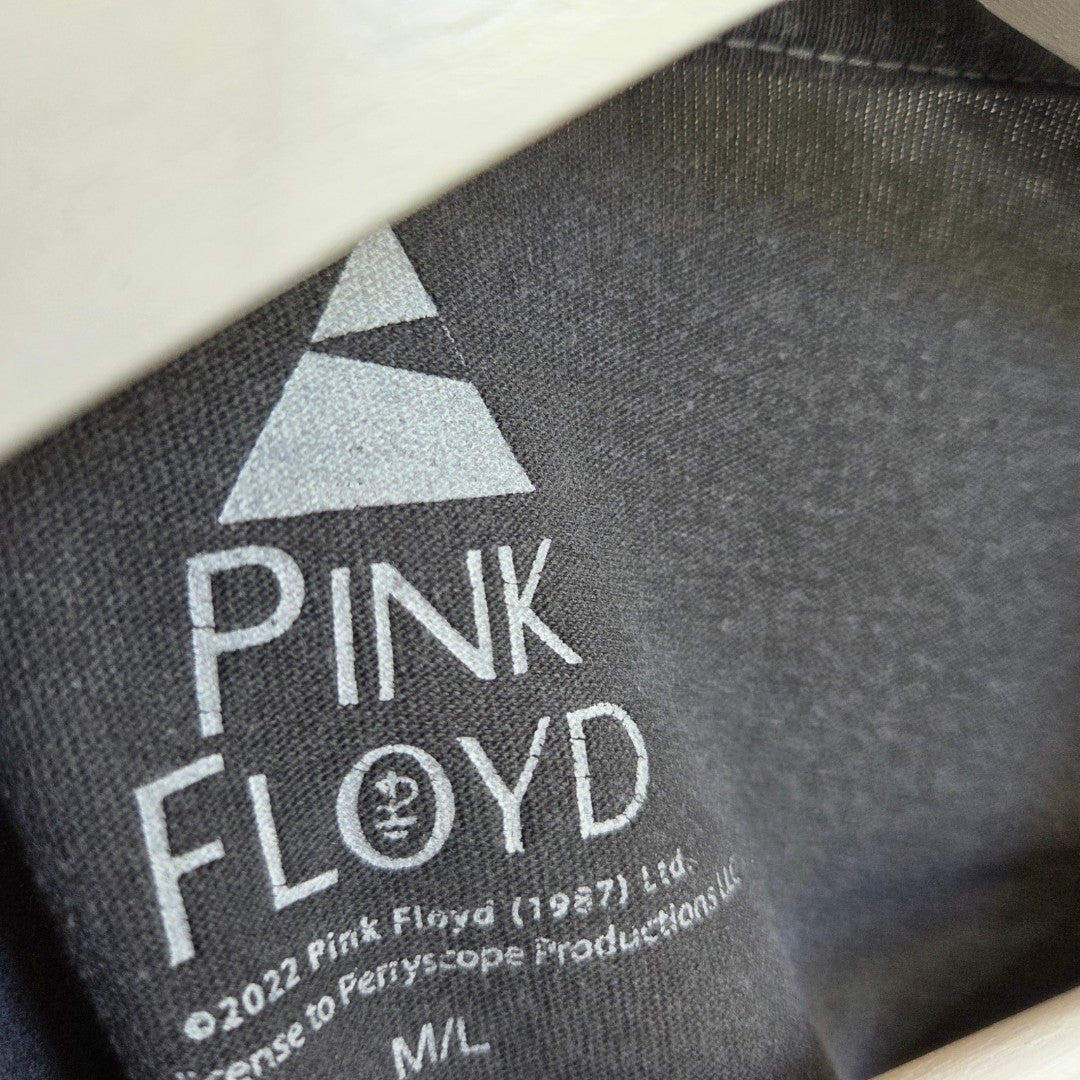 Close-up of a black tag on the Pink Floyd Upcycled Breastfeeding & Pumping Pink/Orange Band t-shirt (M/L), featuring the logo, a triangle above the band name, copyright info, and eco-friendly details as an alternative to typical nursing wear.