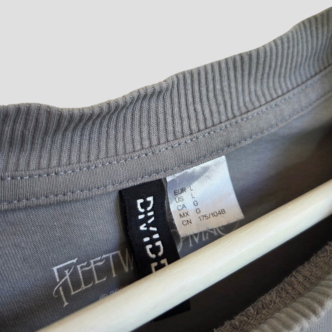 A close-up of an H&M Upcycled Breastfeeding & Pumping Grey/Blue Fleetwood Mac Dreams Band t-shirt (L) shows two tags: a sizing label and a black DIVIDED tag. Made from 100% cotton, making it ideal for comfortable nursing wear.