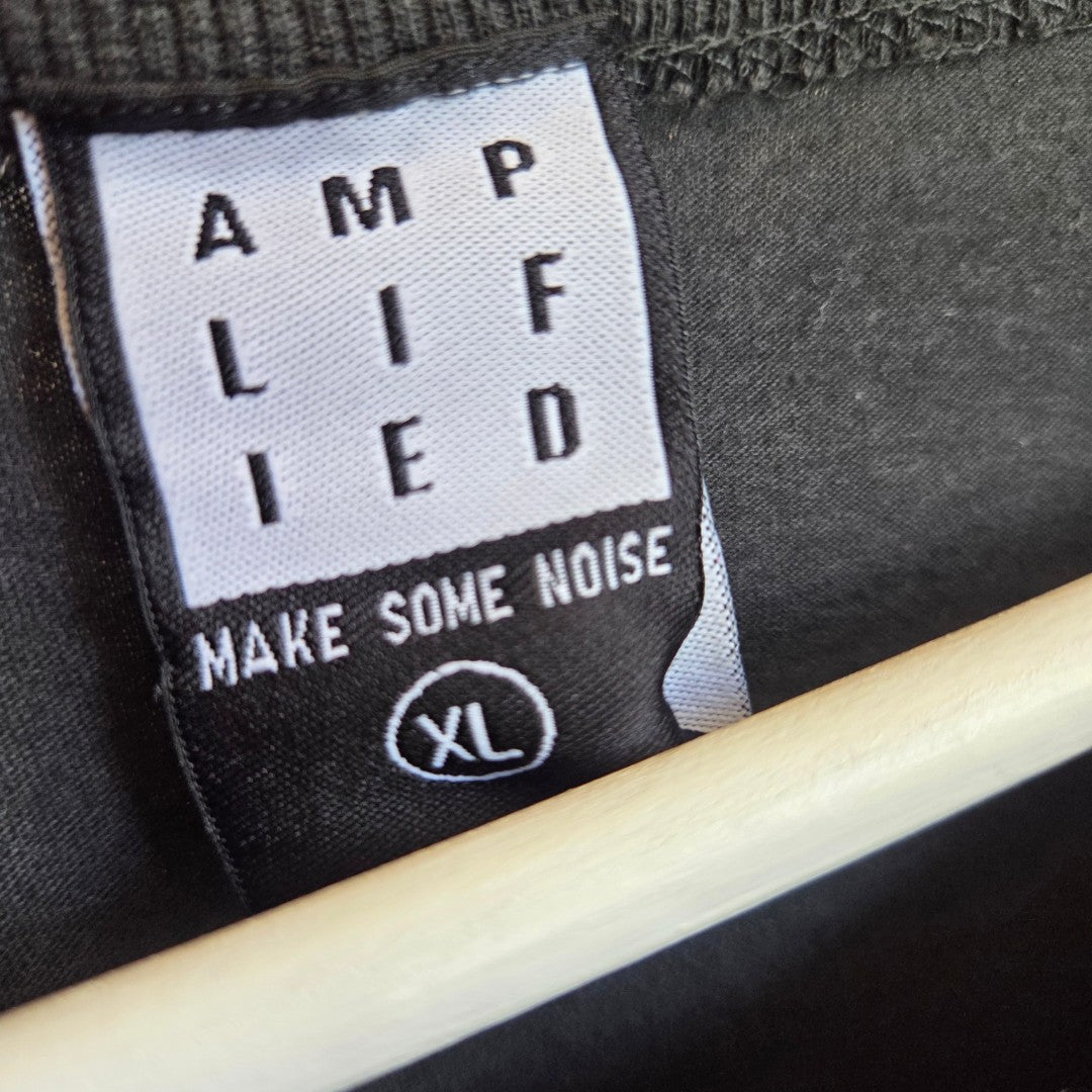 Close-up of a black tag reading "AMPLIFIED" and "MAKE SOME NOISE," with "XL" in a circle, attached to the eco-friendly Upcycled Breastfeeding & Pumping Black/Pink Metallica Band t-shirt XL by Amplified.