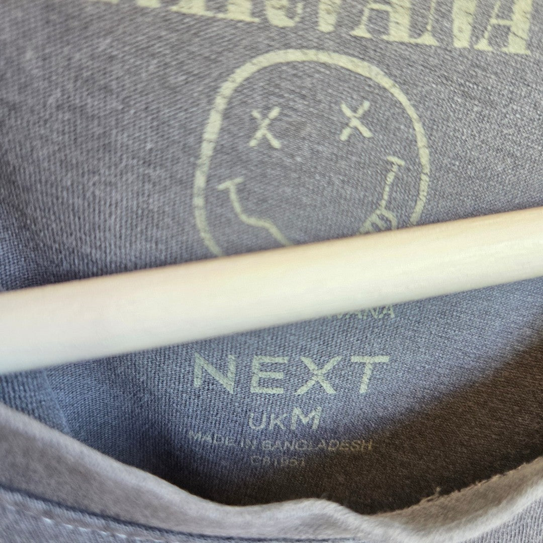 Close-up of an upcycled grey/brown Nirvana band t-shirt (M, NEXT), made in Bangladesh, on a wooden hanger. An eco-friendly, affordable choice for casual breastfeeding or pumping. Logo and label details are visible.