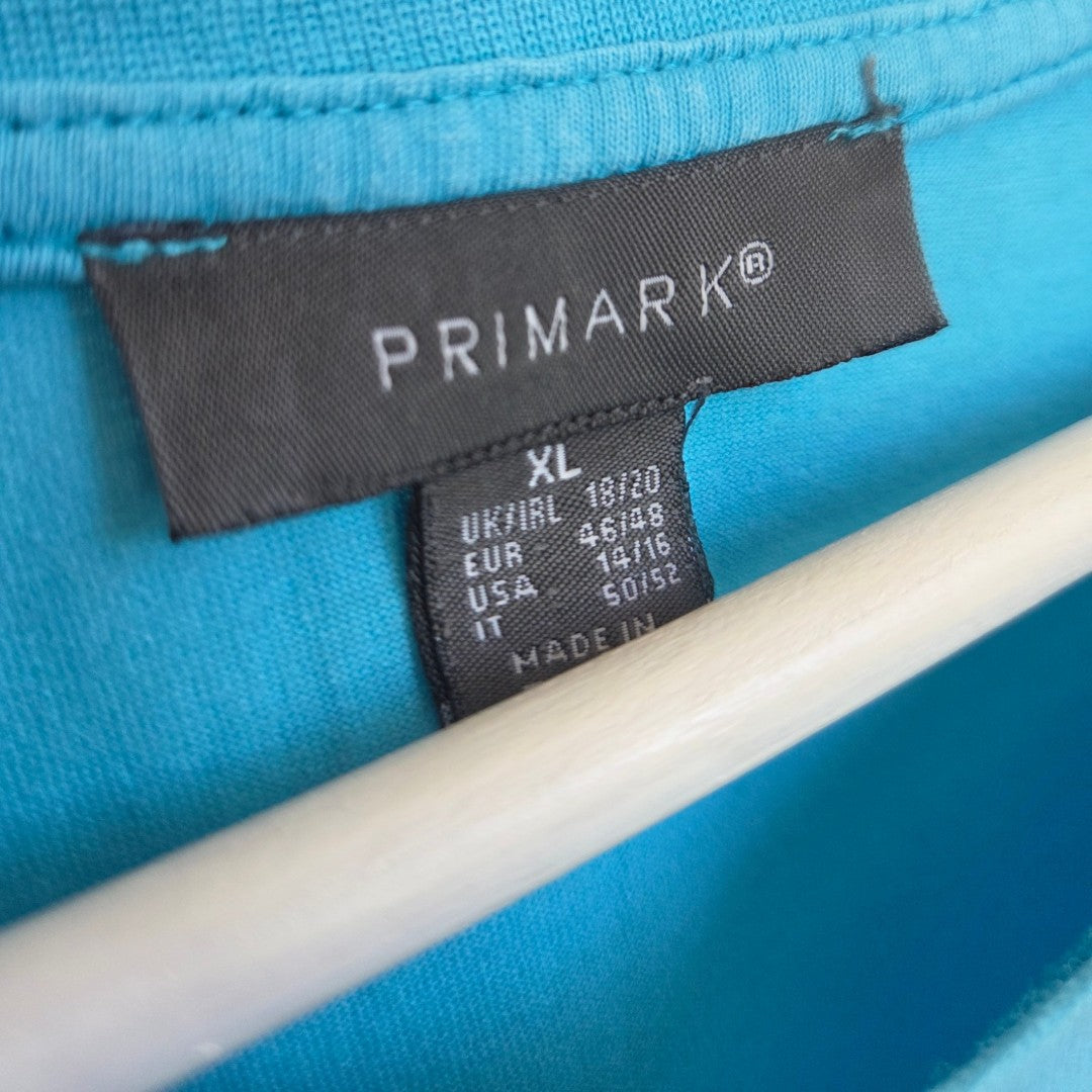 Close-up of a black Primark label on the Upcycled Breastfeeding (Nursing) & Pumping Turquoise/Pink Girl t-shirt in size XL (UK18/20), with partial 'Made in' information visible. Designed for new mothers, this affordable garment hangs on a white hanger against the blue fabric.