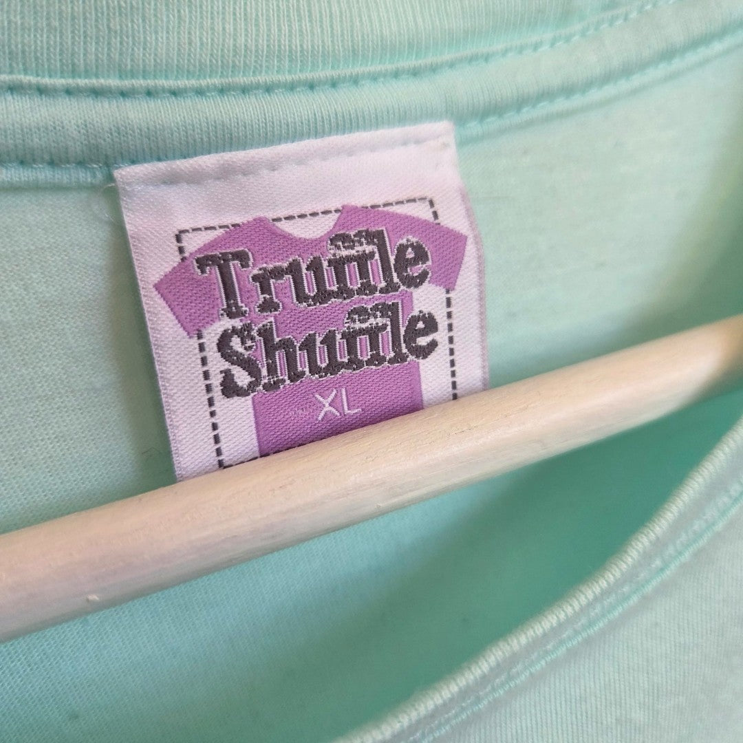 A close-up of a clothing tag on a light blue Upcycled Breastfeeding (Nursing) & Pumping Danny Champion of the World t-shirt in size XL by Truffle Shuffle, designed for those seeking affordable nursing clothes. The tag reads "Truffle Shuffle" with a purple shirt graphic, and a wooden hanger is visible underneath.