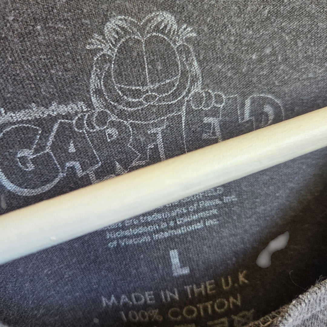 Close-up of a gray label on the Upcycled Breastfeeding (Nursing) & Pumping Garfield Popcorn t-shirt L by Garfield, featuring a cartoon face of Garfield above the word GARFIELD. The label indicates size L, 100% cotton, and MADE IN THE U.K., making it an eco-friendly option for those looking for sustainable fashion. A white hanger is partially visible.