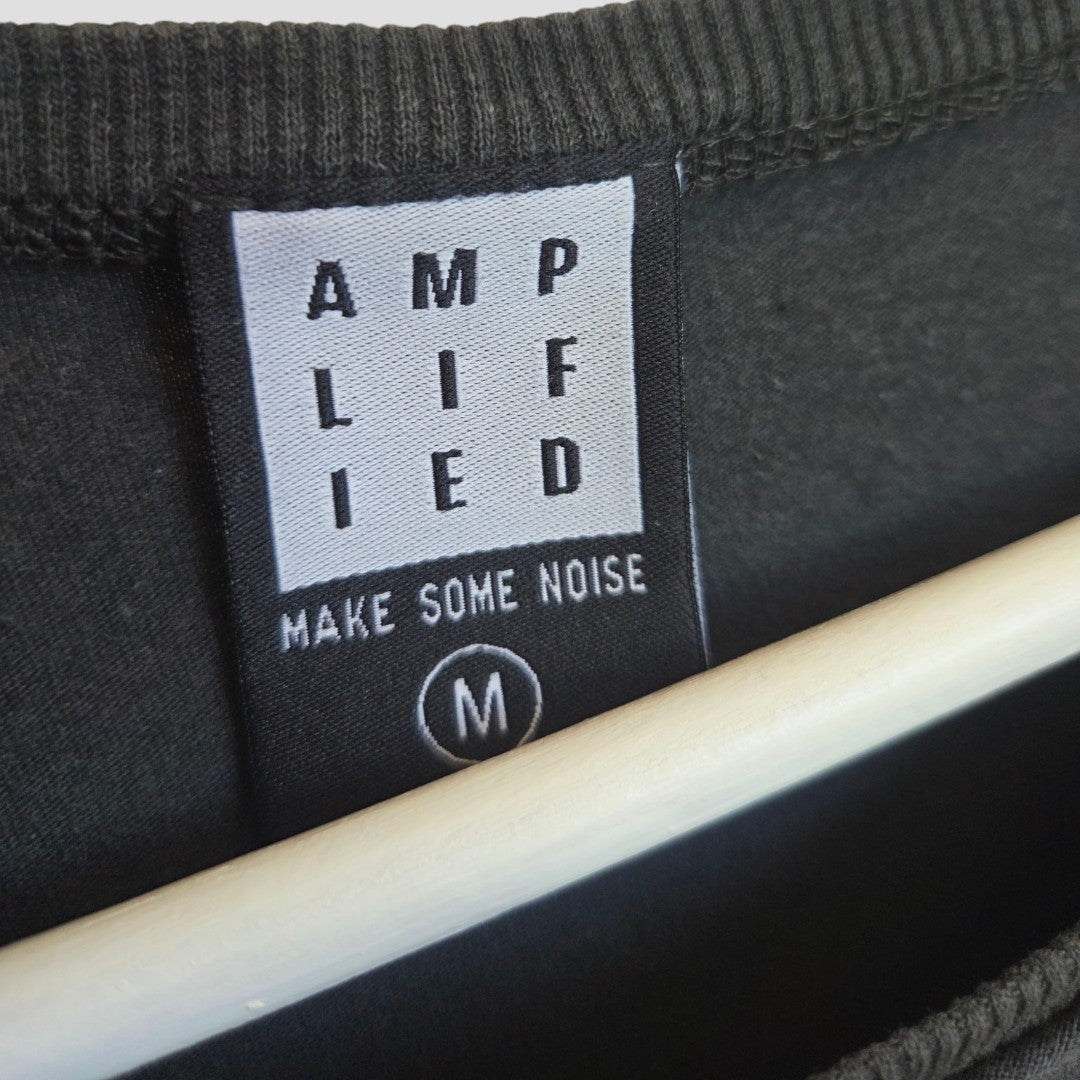 A black clothing label, sewn onto a dark fabric, reads "AMPLIFIED" vertically with "MAKE SOME NOISE" underneath and displays the size M. A white hanger is partially visible—an ideal detail for stylish and sustainable collections such as the Upcycled Breastfeeding (Nursing) & Pumping Black Sabbath Band t-shirt (M) from Amplified.