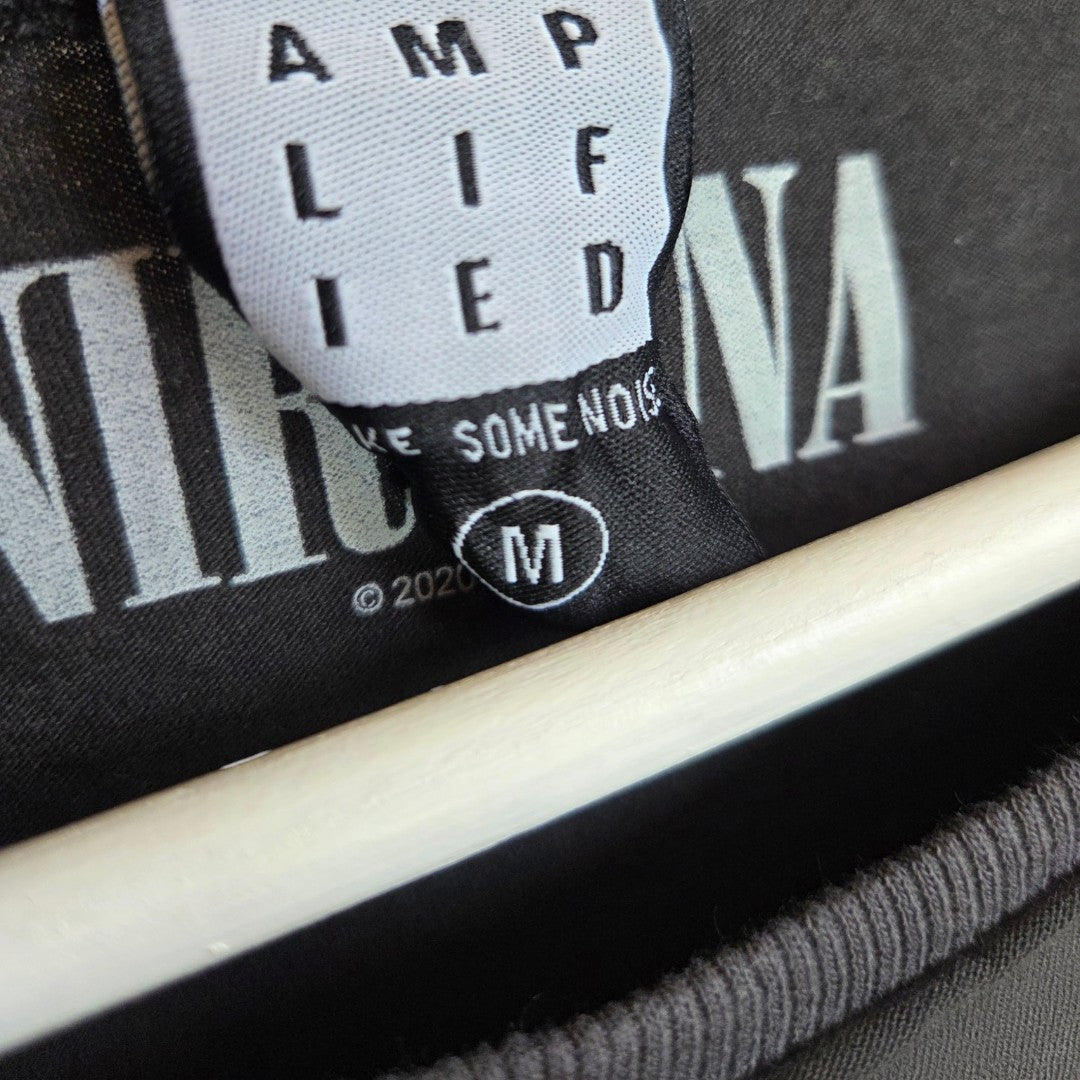 Close-up of a black Amplified clothing tag reading “AMPLIFIED” and “M” for medium, partially covering the Nirvana Snake Band design on an upcycled eco-friendly breastfeeding (nursing) & pumping t-shirt. A wooden hanger is also visible.