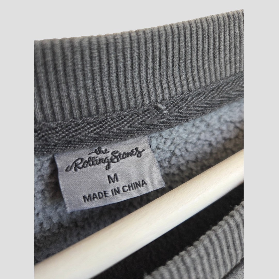 Close-up of a gray clothing tag inside a garment, reading “The Rolling Stones M Made in China,” attached to a ribbed collar with a wooden hanger visible below—highlighting the affordable style of the Primark Upcycled Breastfeeding (Nursing) & Pumping Grey Embroidered The Rolling Stones Band Sweatshirt, size M (100570).