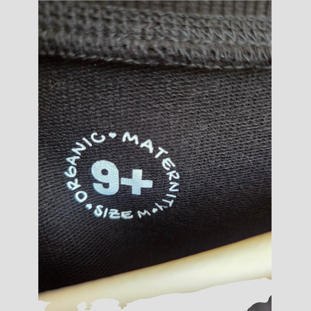 Close-up of a black fabric label with white text reading ORGANIC MATERNITY SIZE M and a large 9+ in the center, highlighting the Nine+ Quarter Upcycled Breastfeeding (Nursing) & Pumping Black Tiger Maternity Sweatshirt (size M, product code 100648) as an ideal choice for comfortable nursing or breastfeeding clothing made from organic cotton.