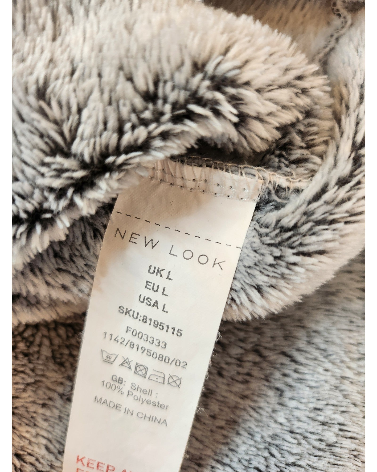 A close-up of a label on soft, gray fabric reveals size info for UK, EU, and USA along with the SKU number. Crafted in China from 100% polyester, this preloved piece by touched out mama is an Upcycled Breastfeeding & Pumping Blanket Hoodie (Size L).