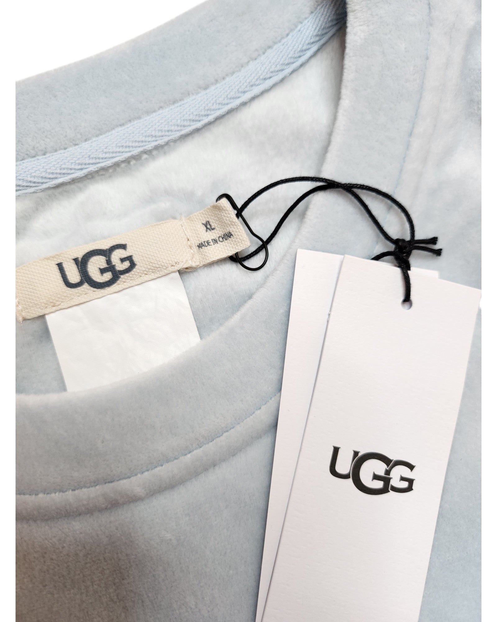A close-up of an XL touched out mama Upcycled Nursing & Pumping UGG t-shirt reveals a soft, light gray garment with a visible collar and logo tag. The eco-friendly piece features a white tag on a black string stating Made in China, combining style, comfort, and sustainability.
