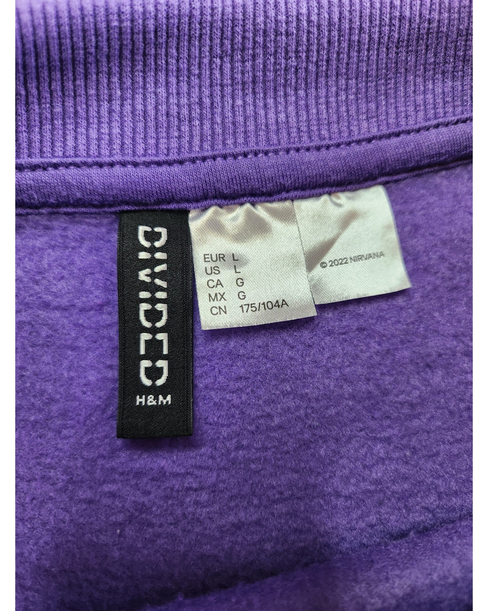 Close-up of clothing labels on a preloved purple fabric feature regional sizing and touched out mama. Another tag reads ©2022 NVIRANA. This upcycled piece, the Upcycled Breastfeeding & Pumping Nirvana Sweatshirt L, showcases a ribbed edge at the top.