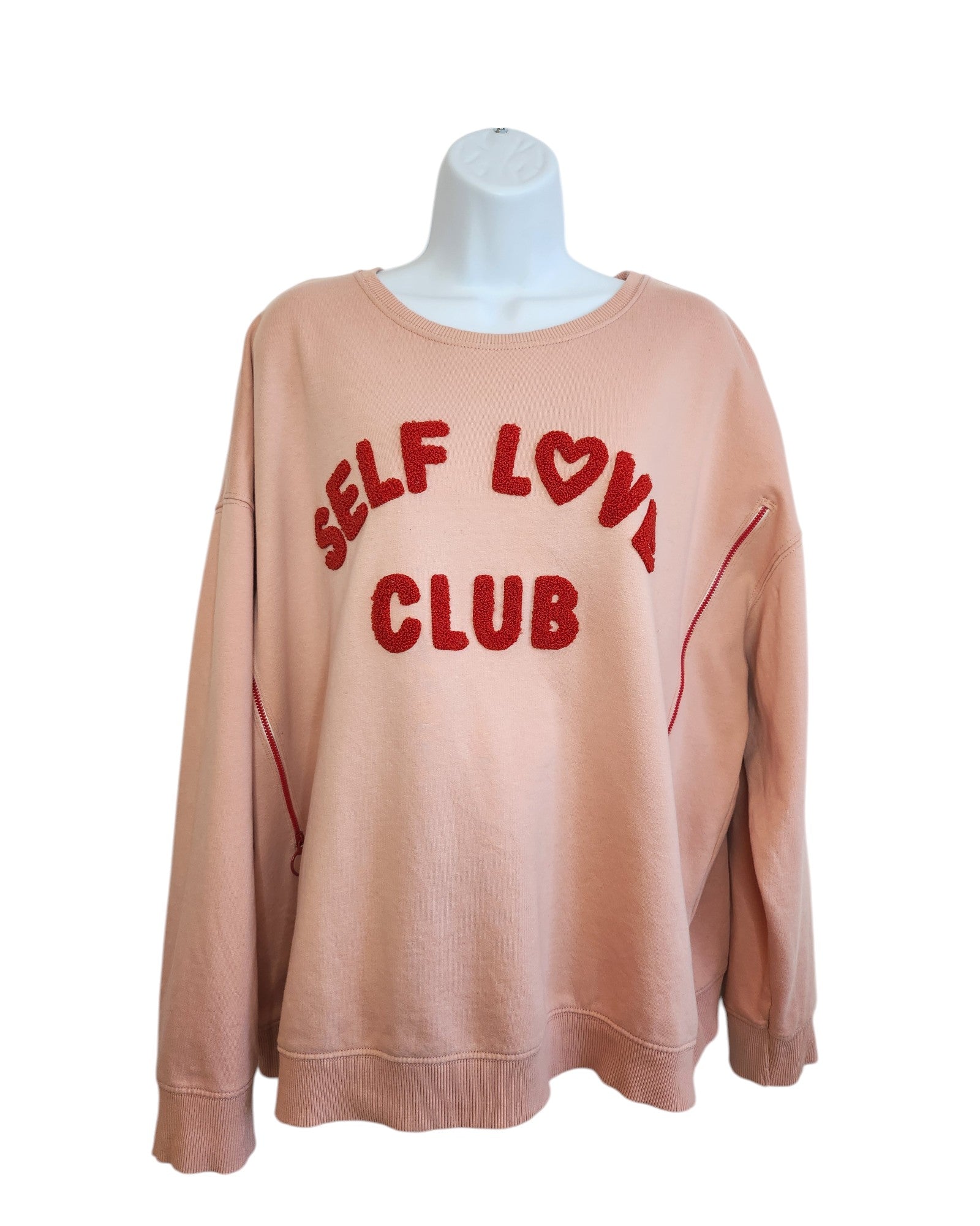 The Self Love Club sweatshirt by touched out mama is a peach-colored, eco-friendly fashion piece with bold red lettering and heart symbol. It features red stitching along the sleeves and sides, perfectly showcased on a white mannequin torso.