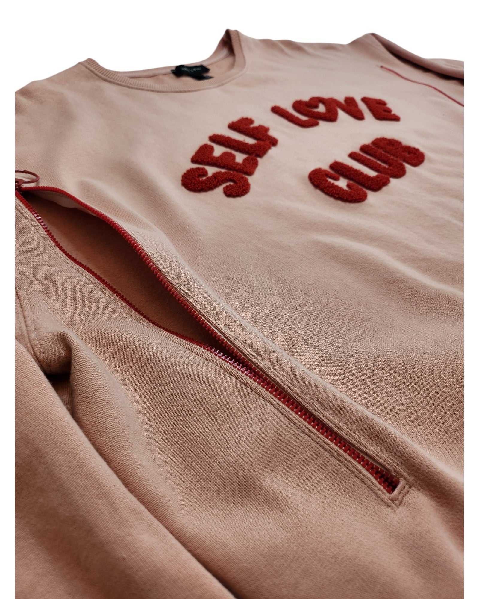 The touched out mama sweatshirt has SELF LOVE CLUB in red textured letters and a diagonal red zipper on soft beige fabric, made from upcycled clothing. The background is white.