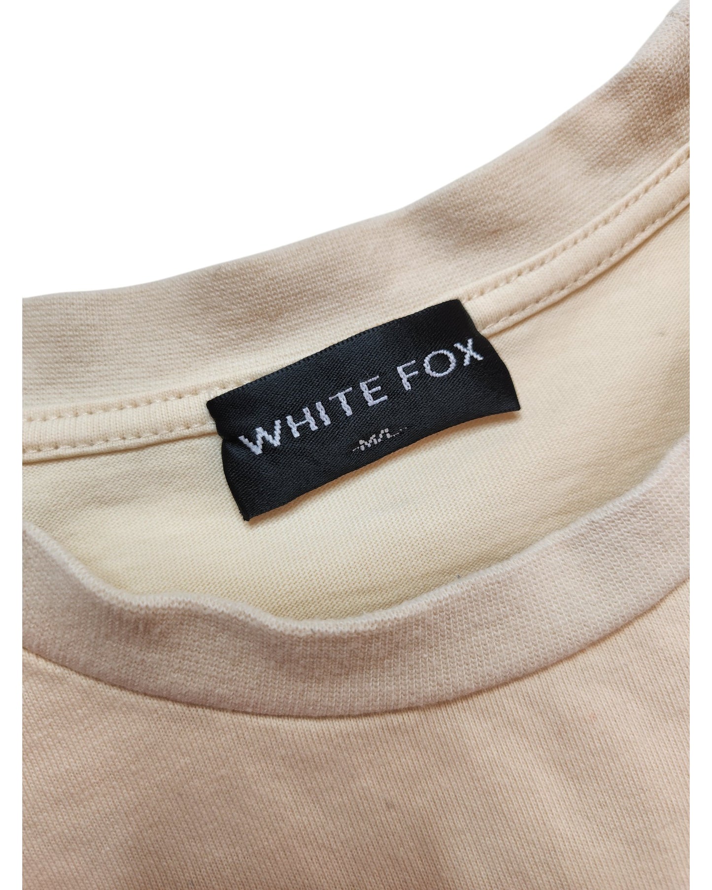 Close-up of the Upcycled Breastfeeding & Pumping White Fox Cream T-shirt M/L by touched out mama, crafted from eco-friendly materials, with a sewn-in black label reading WHITE FOX M/L in white letters. The fabric looks soft and slightly textured.