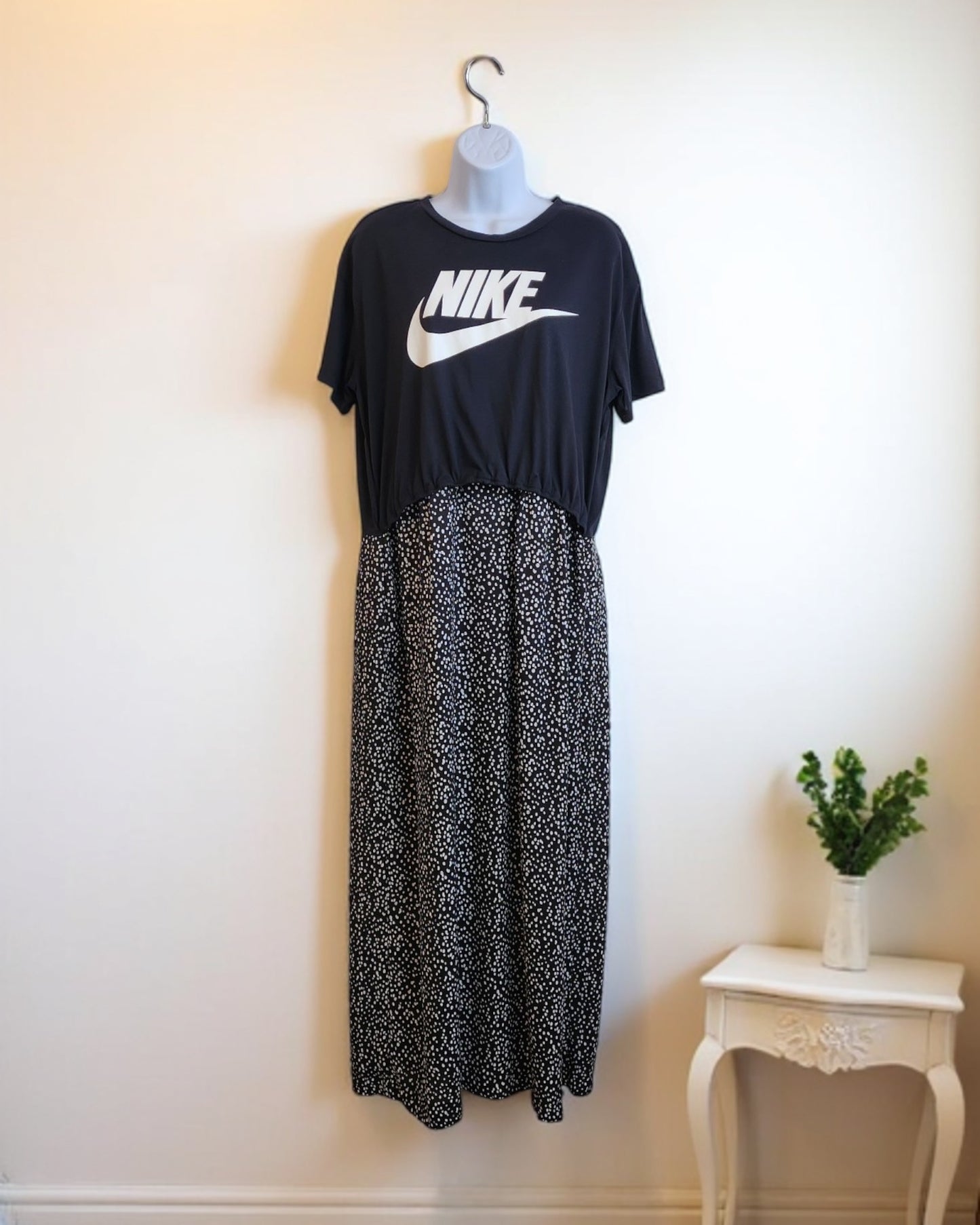 The preloved Upcycled Breastfeeding & Pumping NIKE Maxi Dress M/L from touched out mama, styled over a long black skirt with white dots, is displayed on a mannequin against a cream wall. Its paired with a small white table and green potted plant for an eco-friendly vibe.