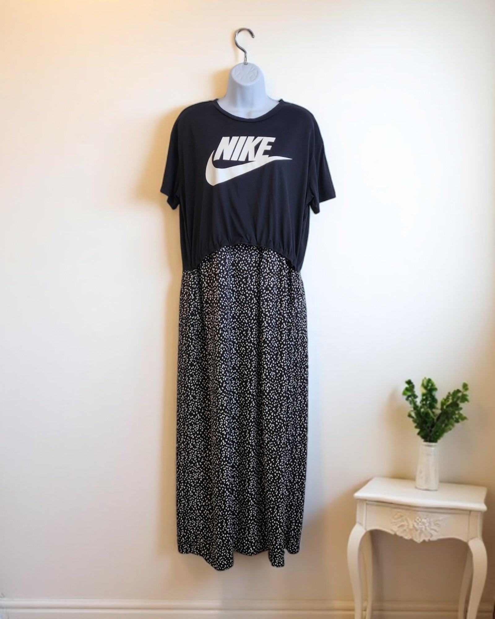 The preloved Upcycled Breastfeeding & Pumping NIKE Maxi Dress M/L from touched out mama, styled over a long black skirt with white dots, is displayed on a mannequin against a cream wall. Its paired with a small white table and green potted plant for an eco-friendly vibe.