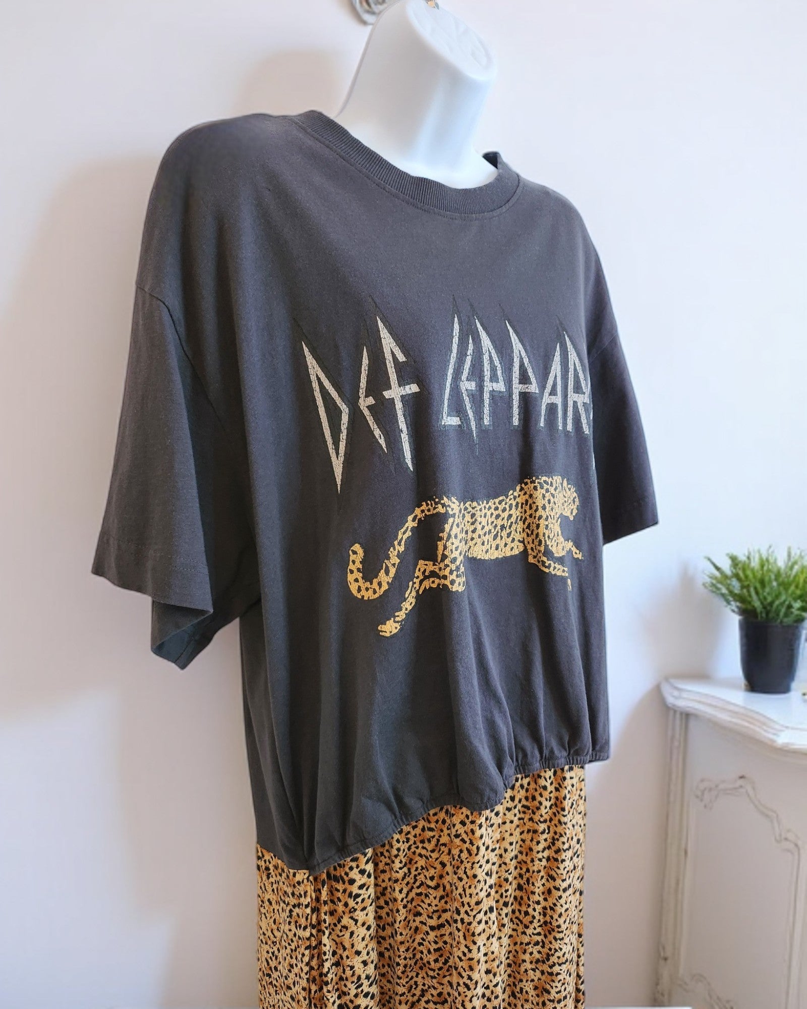 The touched out mama Upcycled Breastfeeding & Pumping Def Leppard Maxi Dress M/L takes center stage with its vibrant design, styled alongside a preloved leopard print skirt on a mannequin. In the background, a small potted plant adds sustainable charm to the scene.