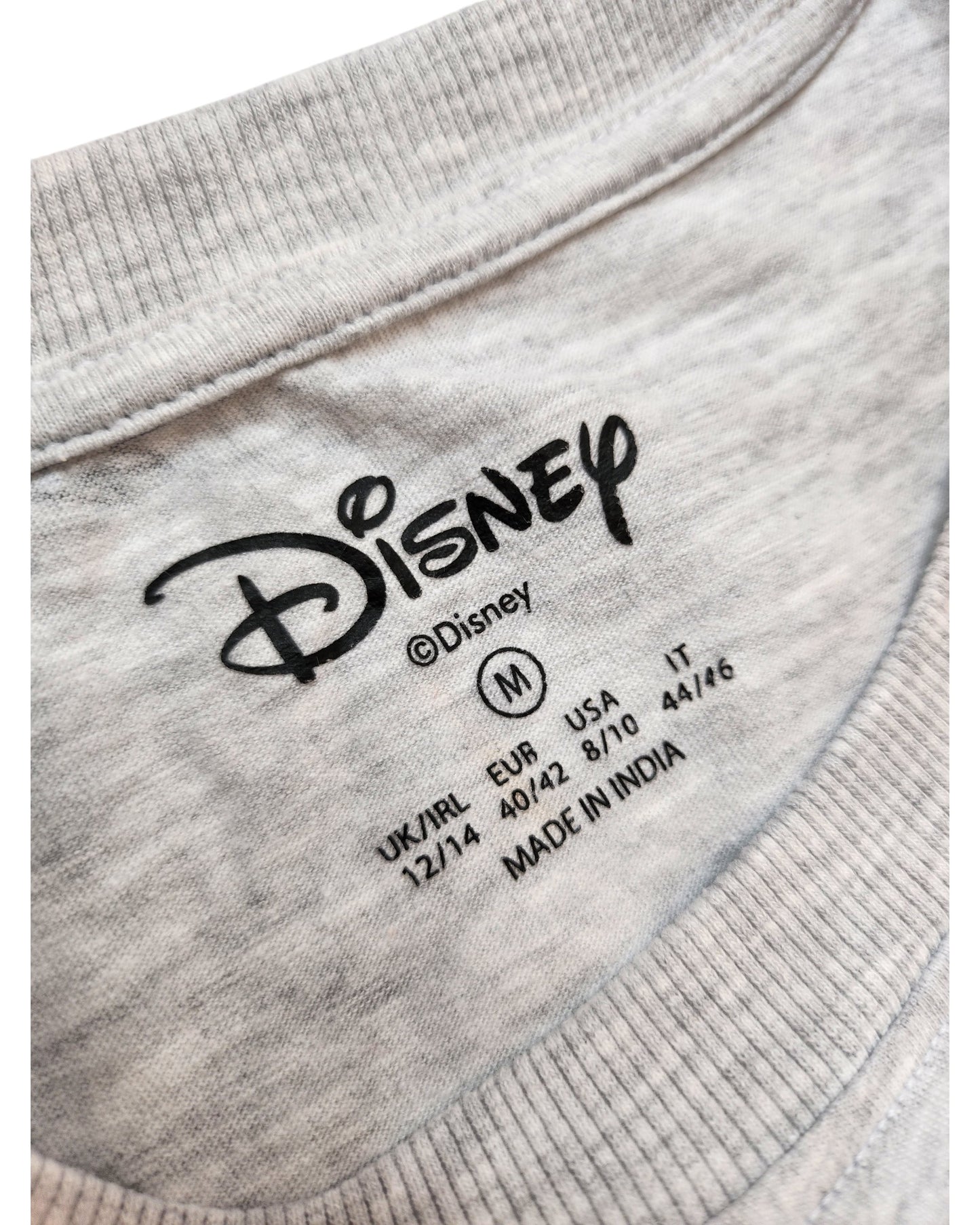 A close-up of a gray touched out mama Disney t-shirt label shows size medium (M) for UK/IRL, EUR, USA, and IT. This upcycled nursing & pumping Stitch tee is crafted in India, blending style and sustainability.